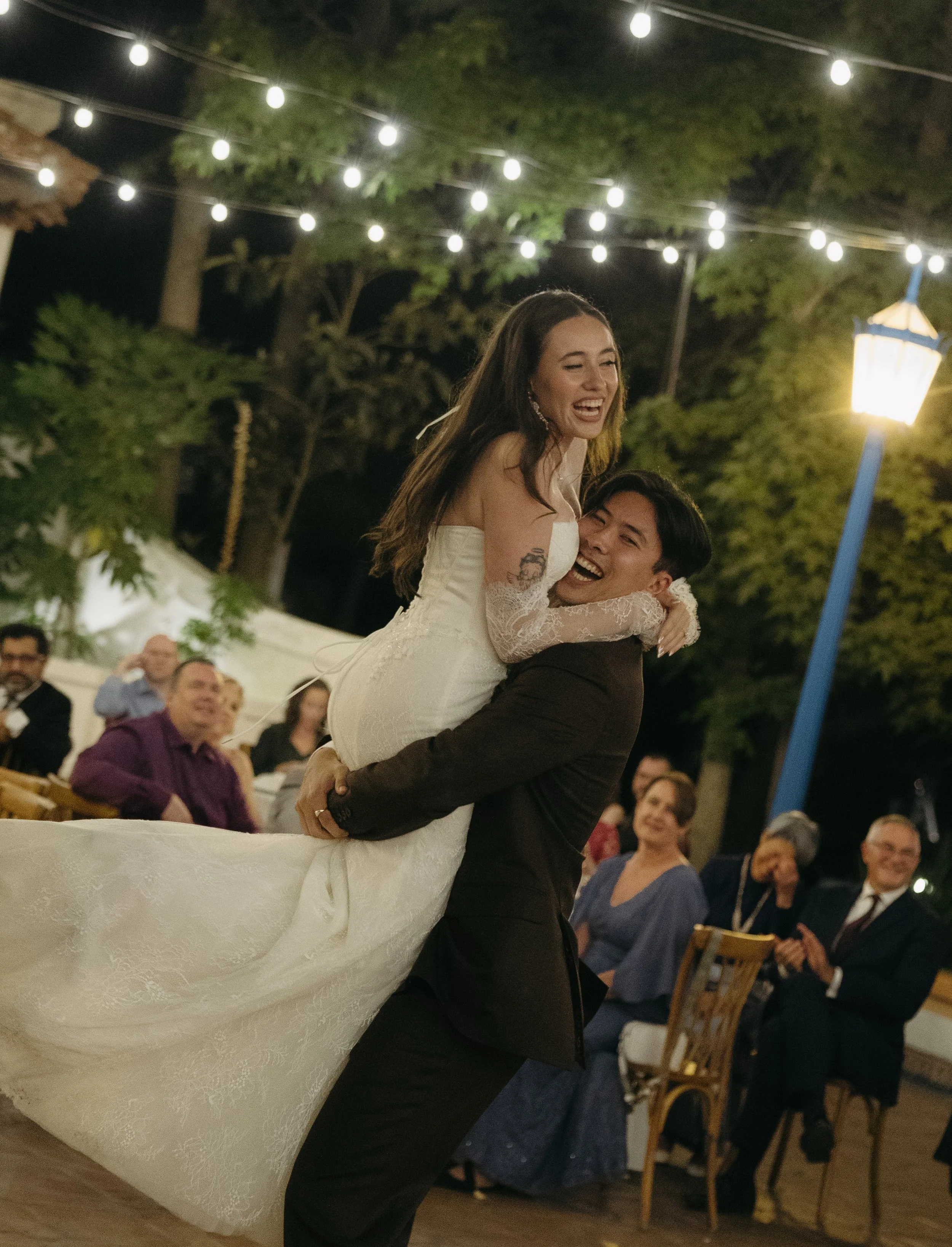 Bride in a white wedding dress being lifted and celebrated by groom in a tuxedo during a nighttime outdoor wedding reception with string lights and guests in the background.