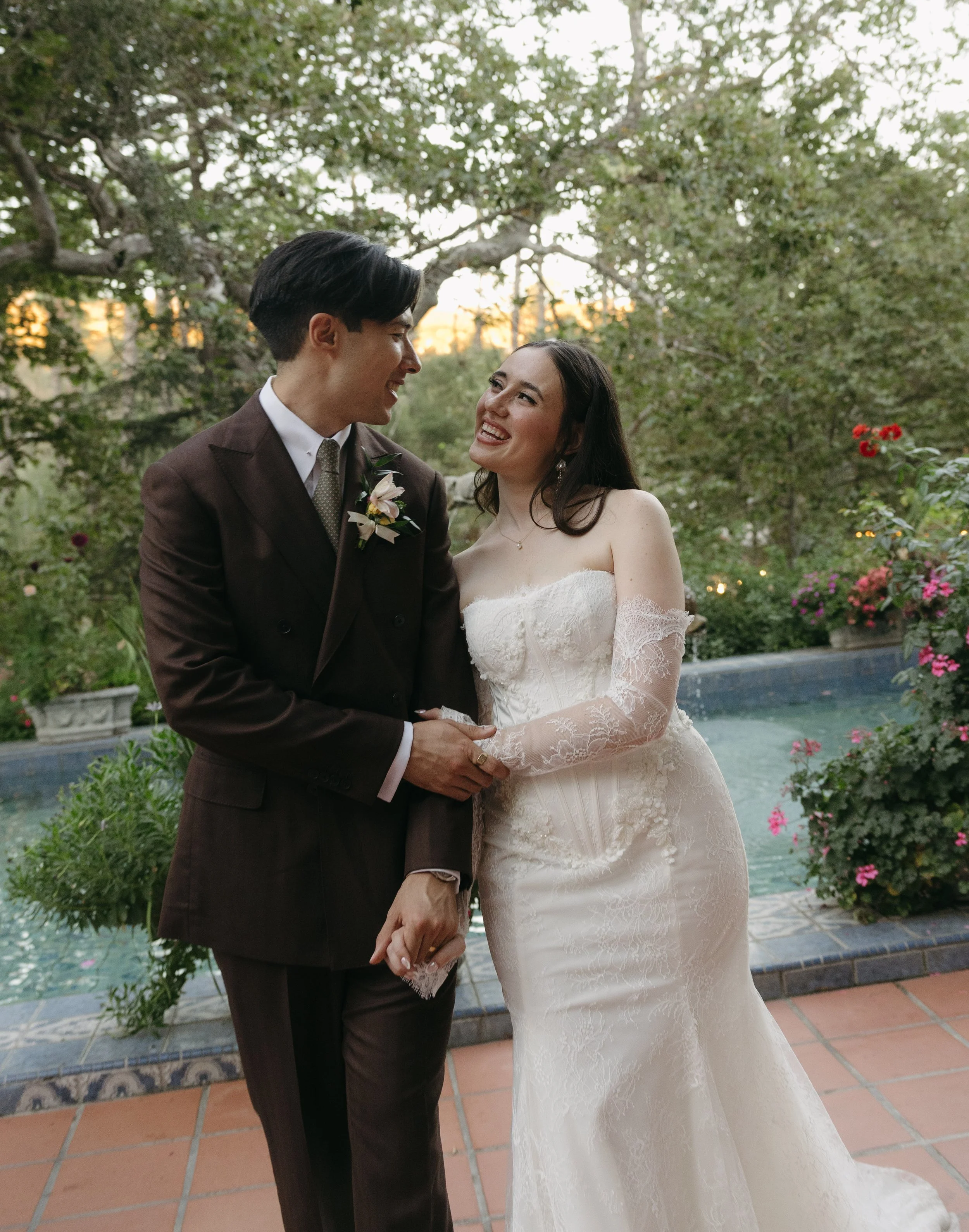 A newly married couple holding hands and smiling at each other outdoors during sunset, with a garden and pool in the background of a Rancho Las Lomas Wedding in Los Angeles