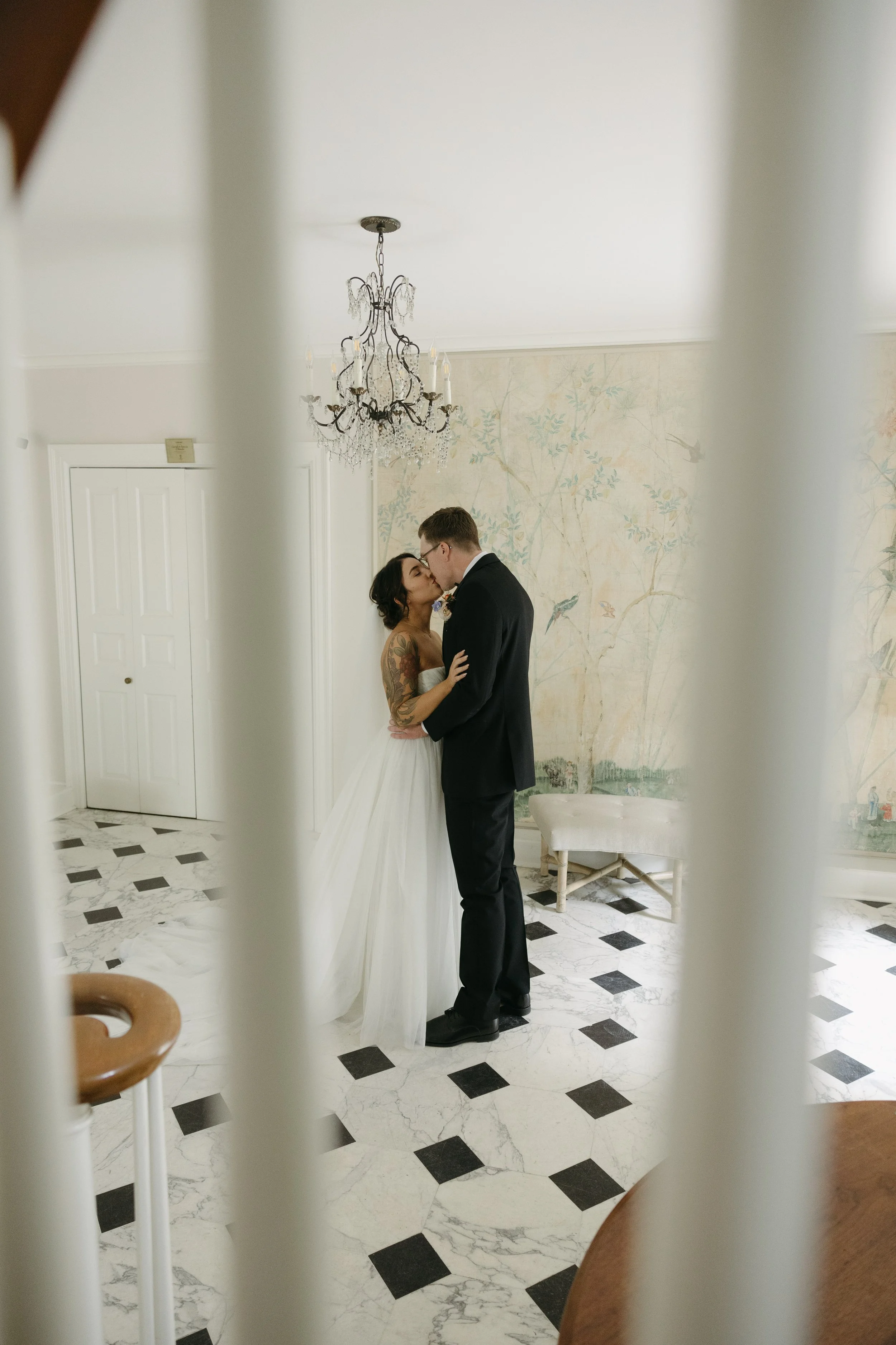 A bride and groom sharing a kiss during their wedding, seen through white vertical bars in an elegant room with a chandelier, patterned wallpaper, and a black and white marble floor.
