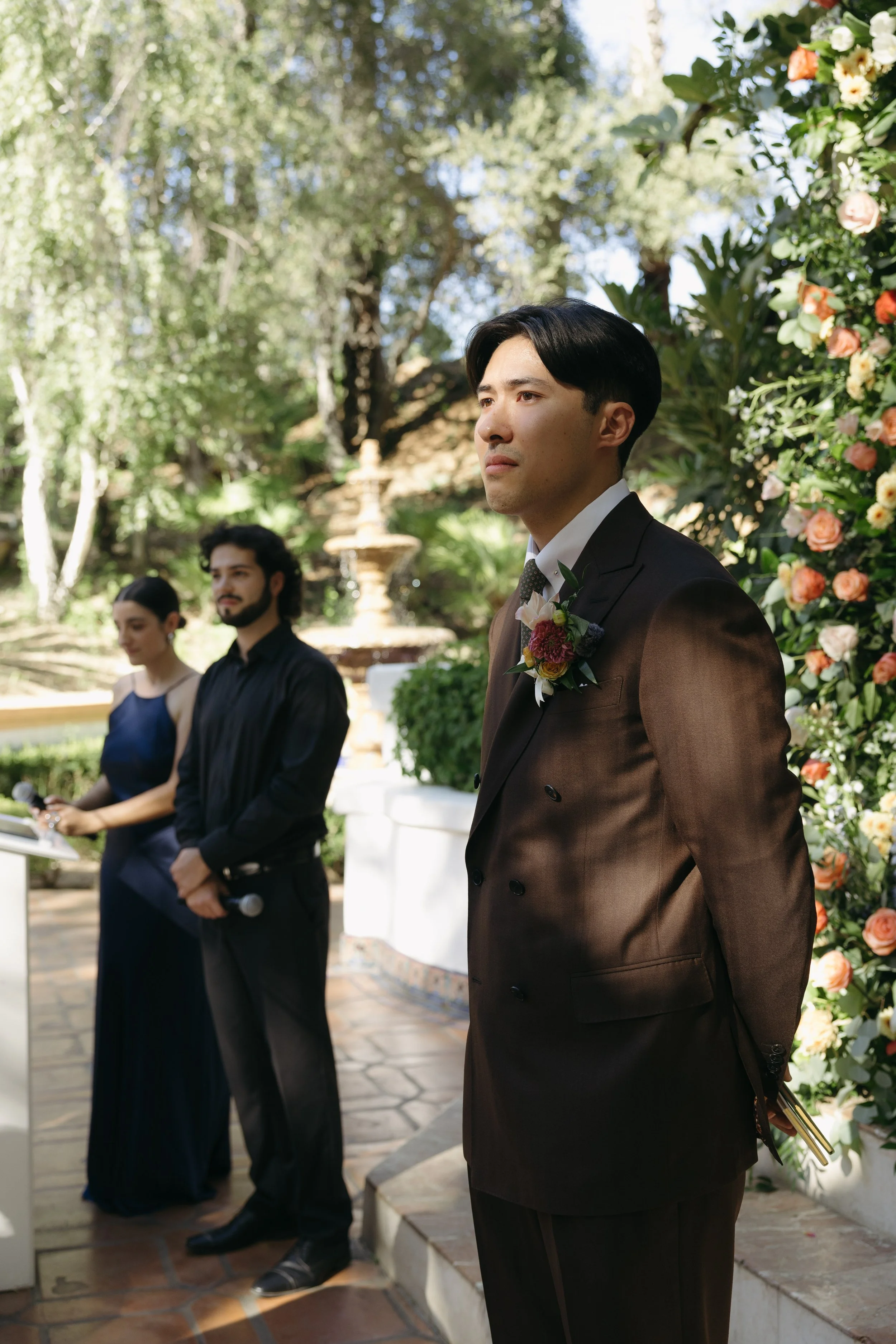 A groom in a brown suit with a boutonniere stands in front of a floral arch at an outdoor wedding ceremony, with a woman and a man in dark clothing in the background.