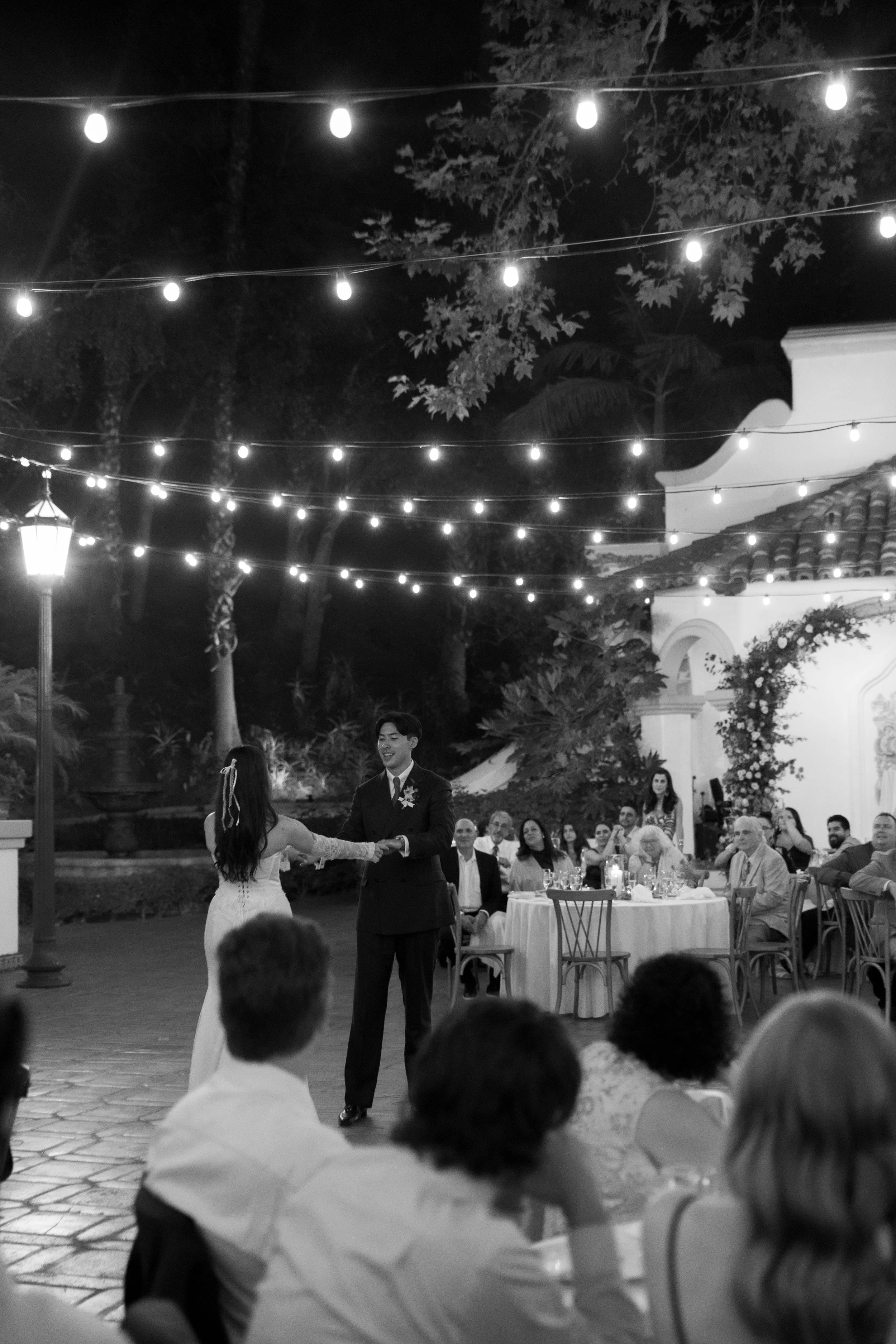 A black and white photo of a wedding reception outdoors at night, with string lights overhead. The bride and groom are dancing in the center, surrounded by seated guests.