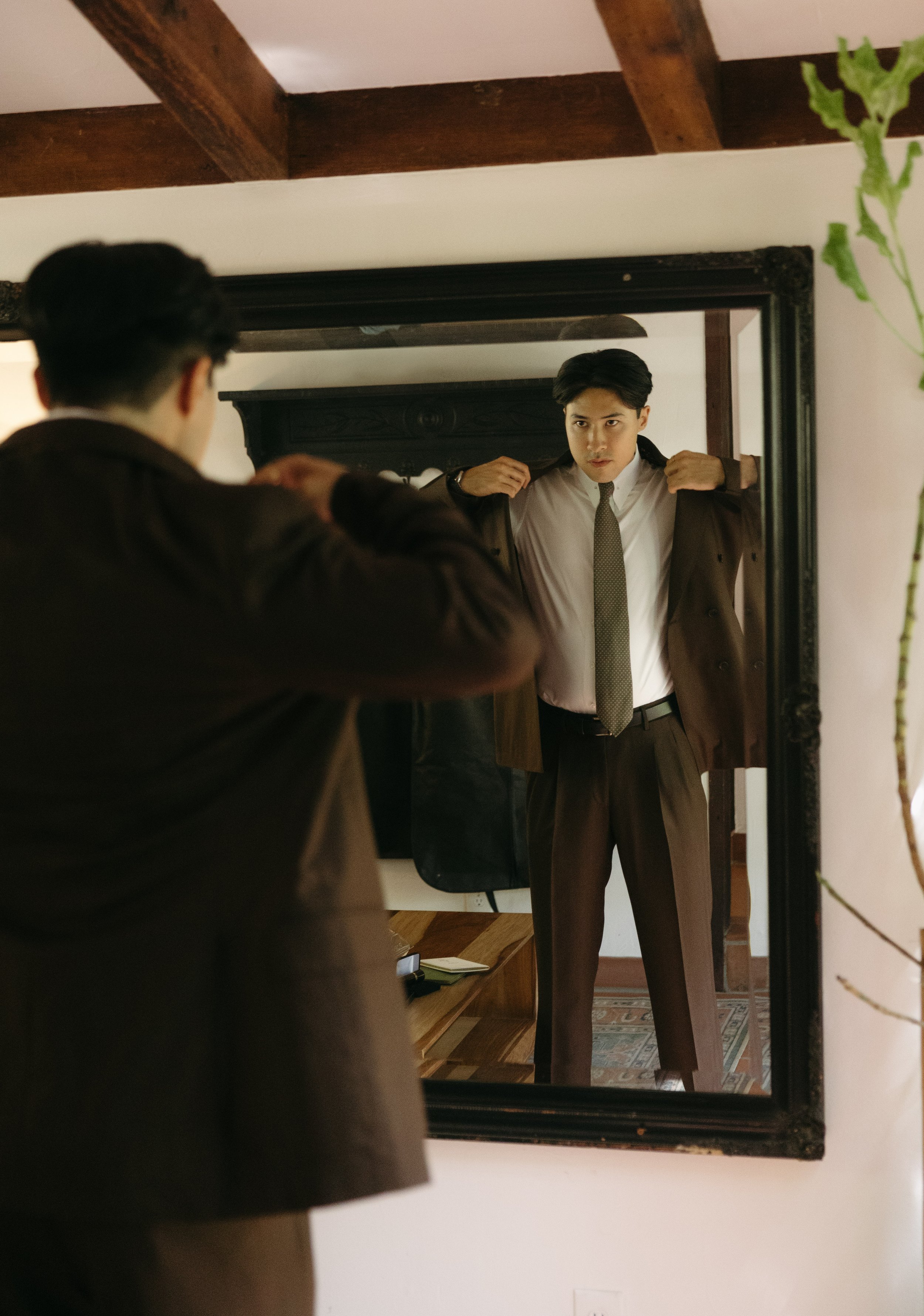 A groom is putting on his jacket in front of a mirror. His reflection shows him adjusting his jacket collar, and a wooden cabinet and some household items are visible in the background at a Rancho Las Lomas Wedding in Los Angeles