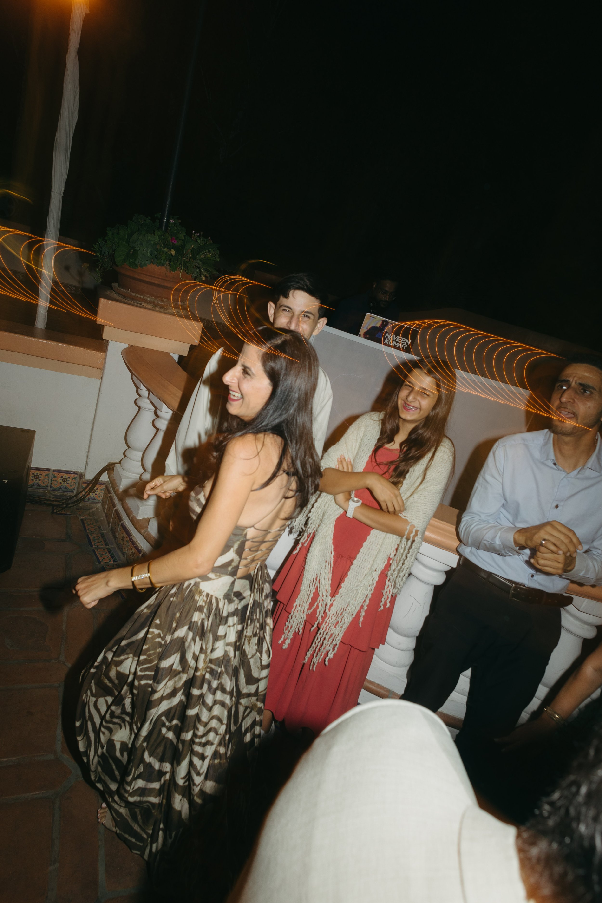 A group of people dancing and socializing at a lively indoor or outdoor party with colorful lighting and a DJ in the background.