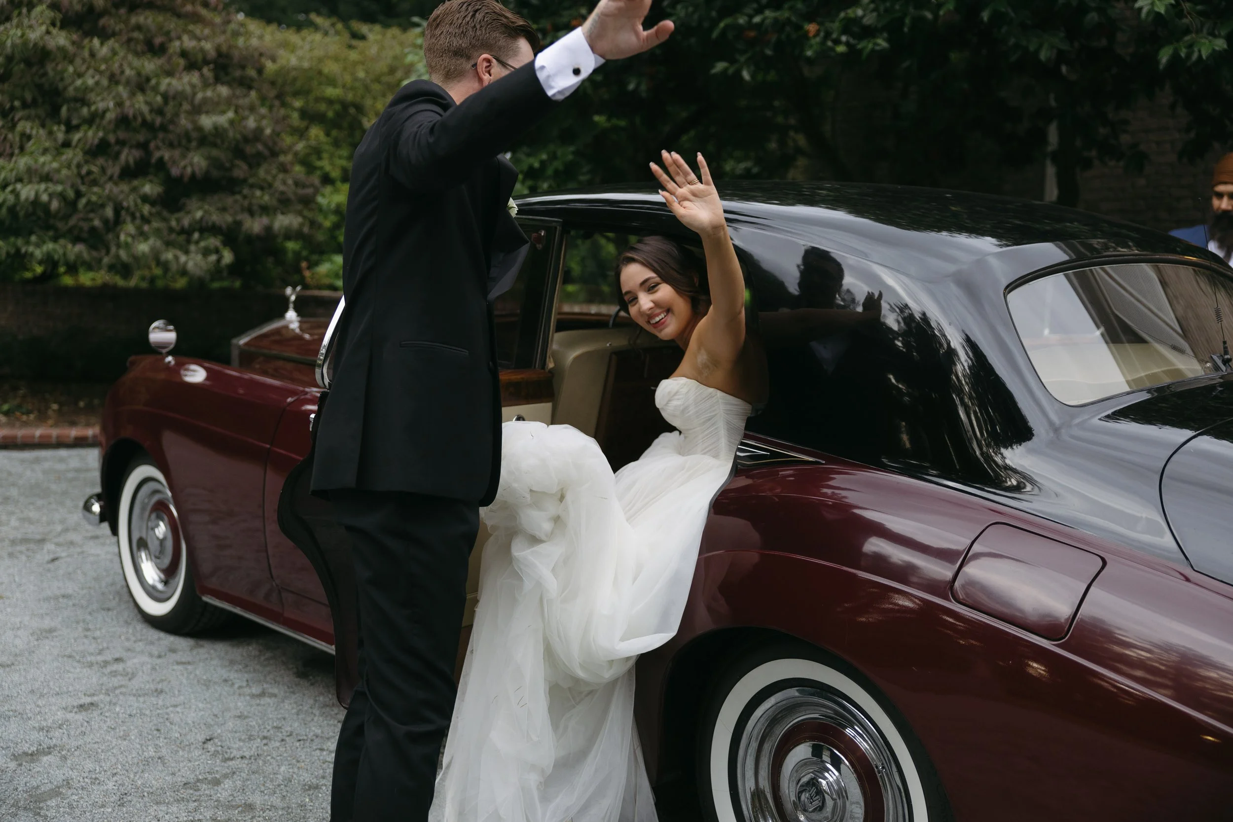 Bride in a white wedding dress smiling and waving from a vintage maroon and black car, with a man in a tuxedo standing outside the car waving back at a European inspired Lakewold Gardens wedding in Seattle
