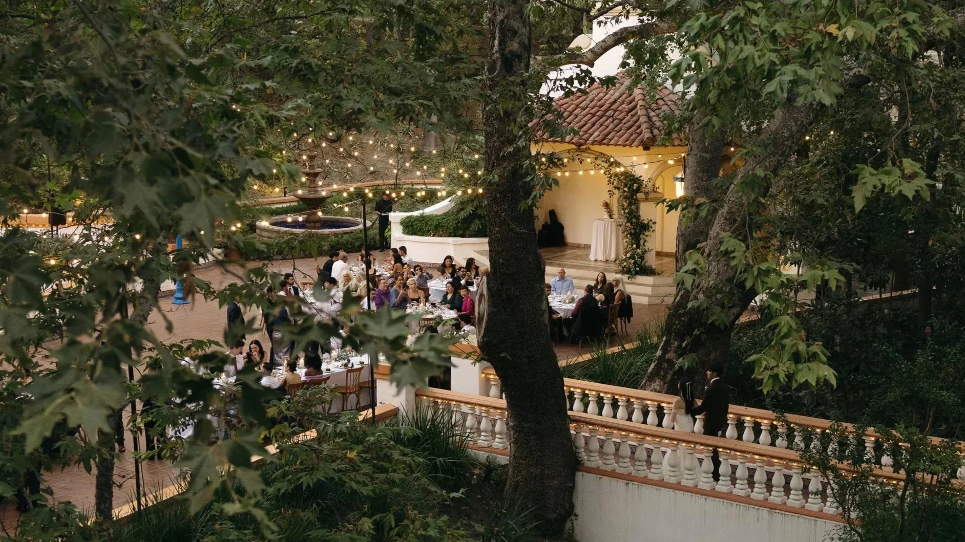 Guests dining at outdoor tables with string lights overhead, surrounded by trees and a decorative building with a red tiled roof at an outdoor wedding reception at Rancho Las Lomas in los Angeles.