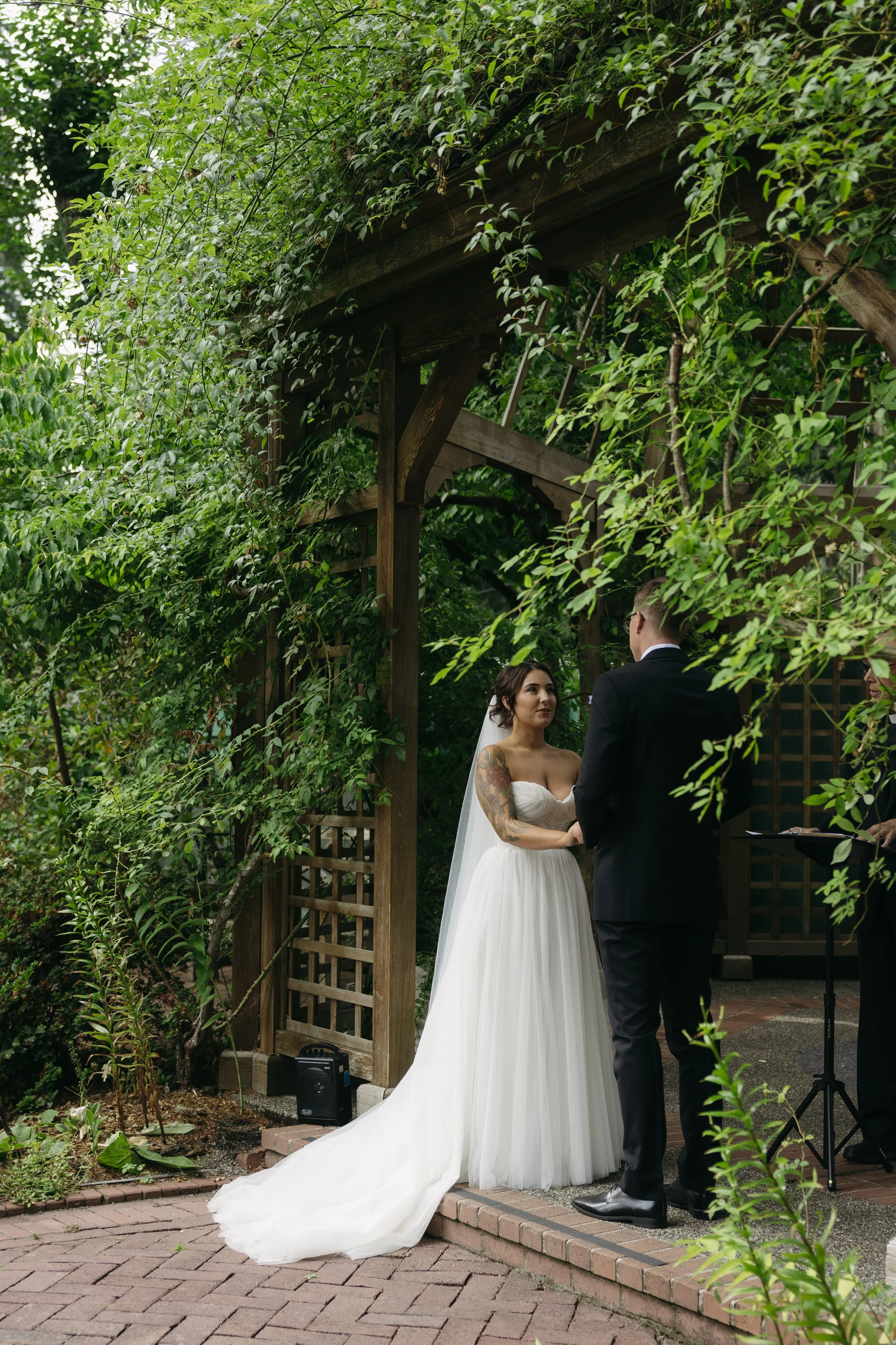 A bride and groom exchanging vows outdoors in a lush, green garden. The bride is wearing a strapless white wedding gown with a long train and veil, and has tattoos on her arms. The groom is dressed in a black suit. An officiant stands to the right, h
