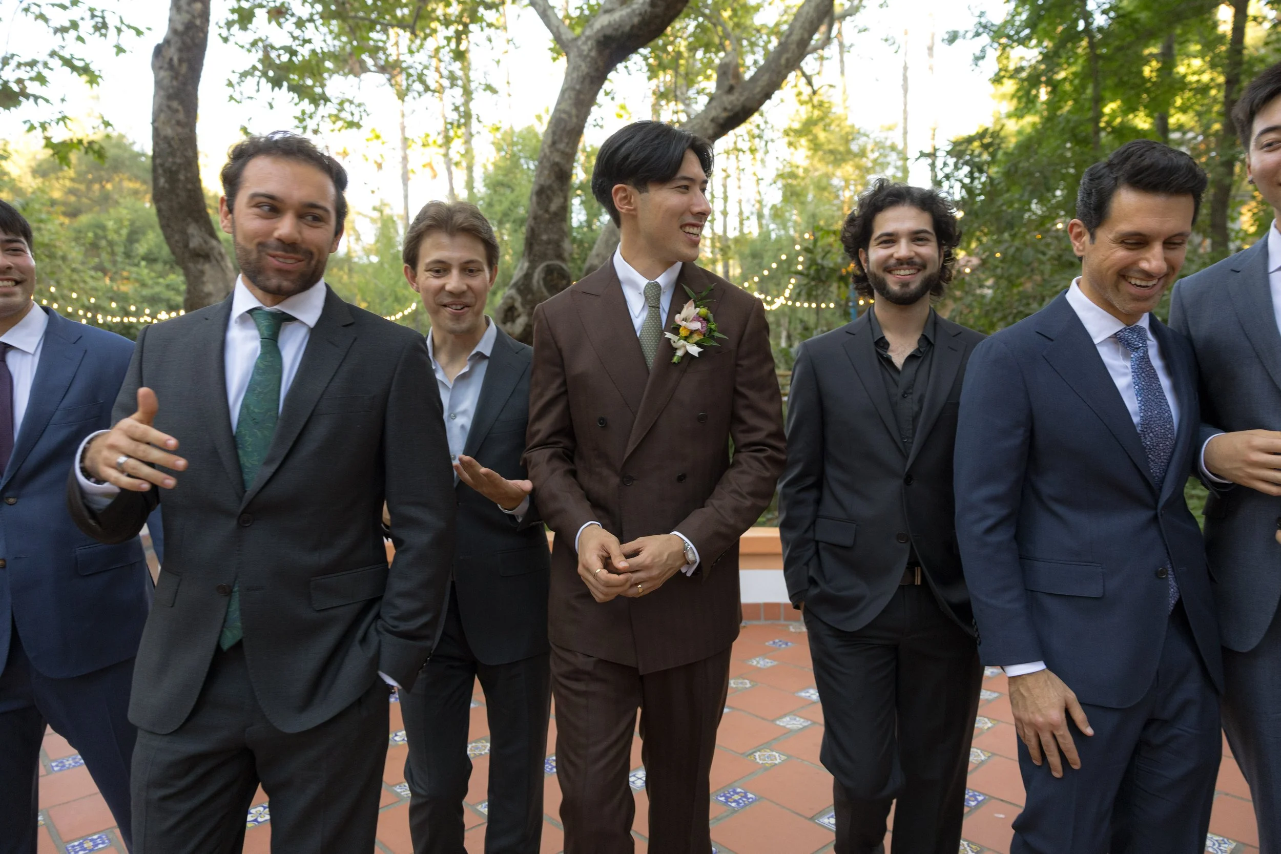 Group of men dressed in suits at an outdoor event with string lights, trees, and tiled flooring in the background.
