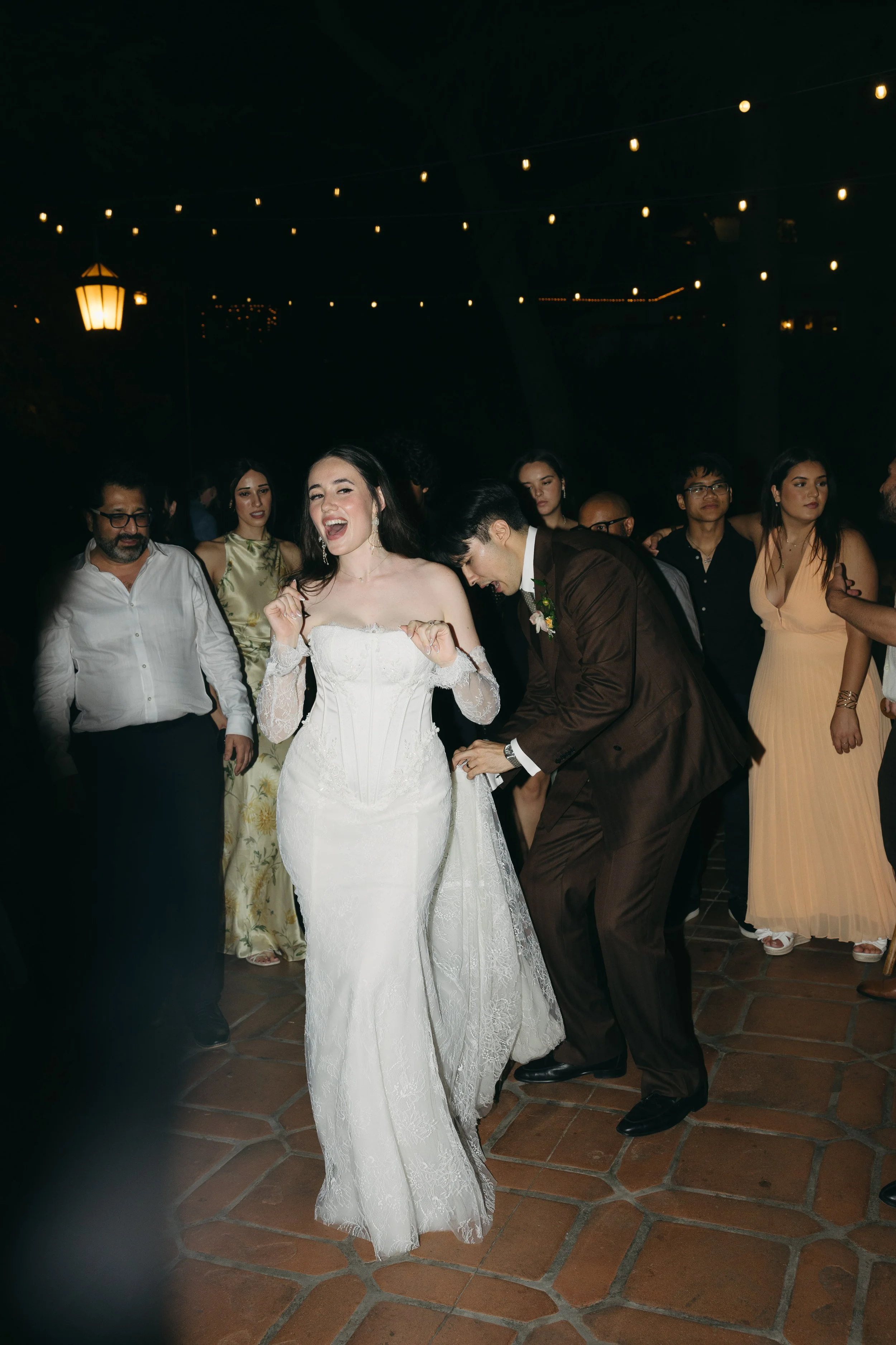 Couple dancing at a wedding reception, with the bride in a white wedding gown and the groom in a brown suit, surrounded by guests under string lights at night.