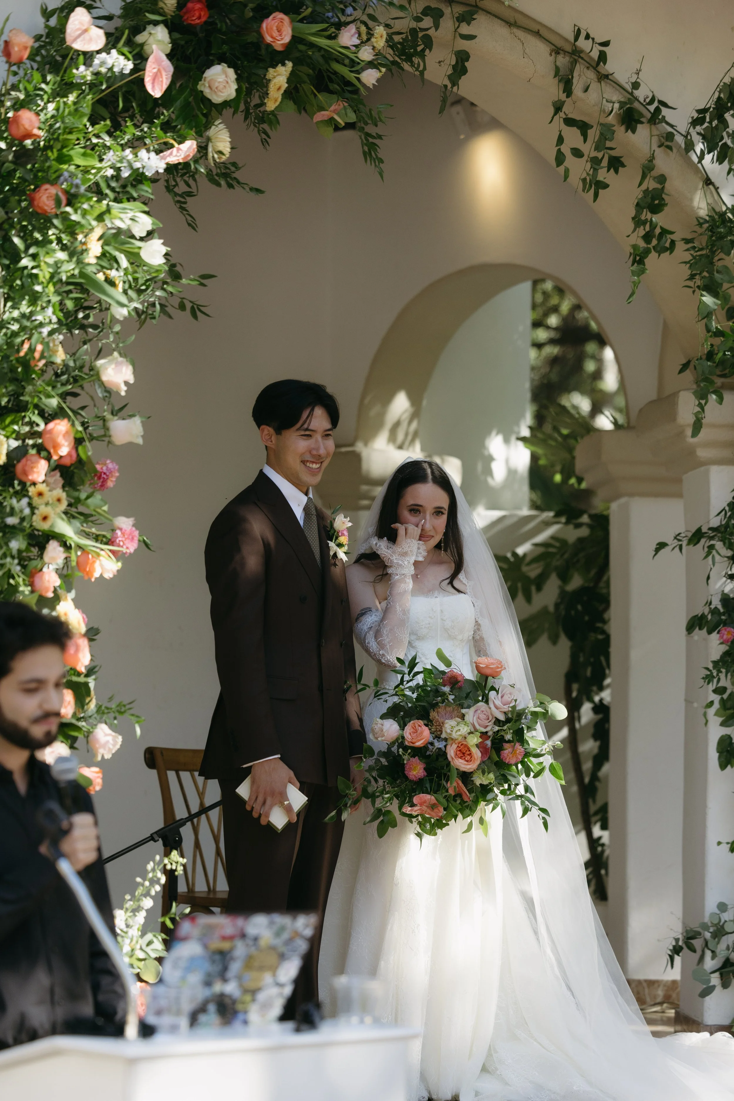 A bride and groom standing together during a wedding ceremony surrounded by colorful flowers and greenery, with a person speaking into a microphone in the foreground at a Rancho Las Lomas Wedding in Los Angeles