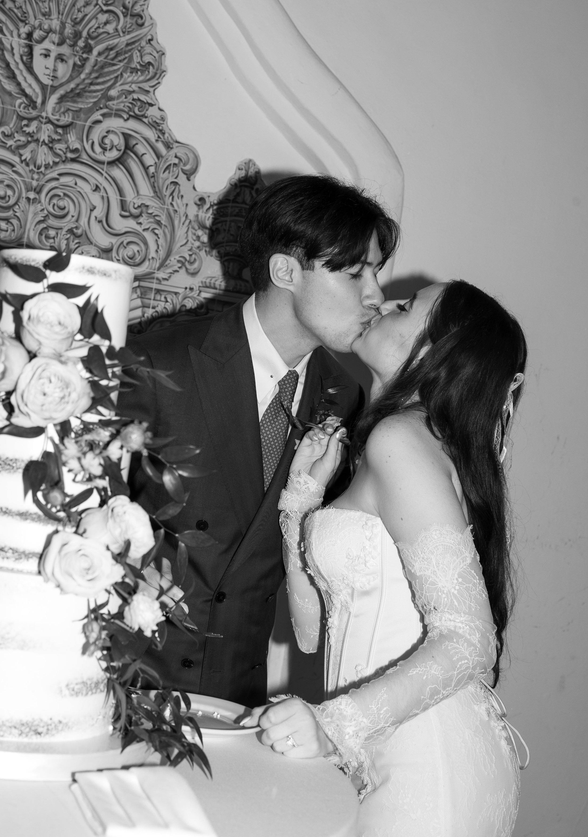 A couple in wedding attire sharing a kiss at their wedding reception, with a wedding cake and floral arrangement in the foreground.