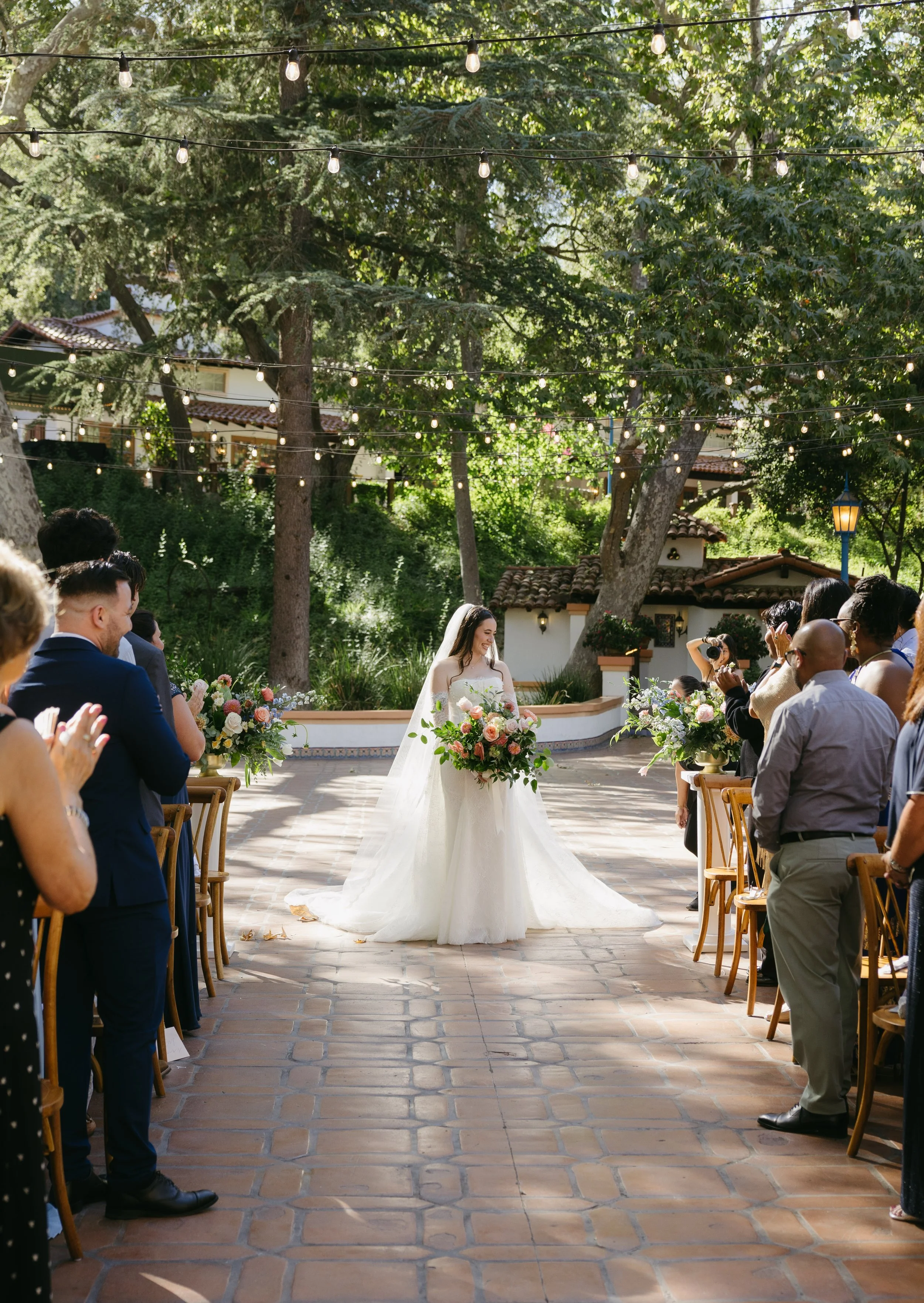 A bride holding a bouquet in an outdoor wedding ceremony surrounded by seated guests, with string lights overhead and lush green trees in the background at a Rancho Las Lomas Wedding in Los Angeles
