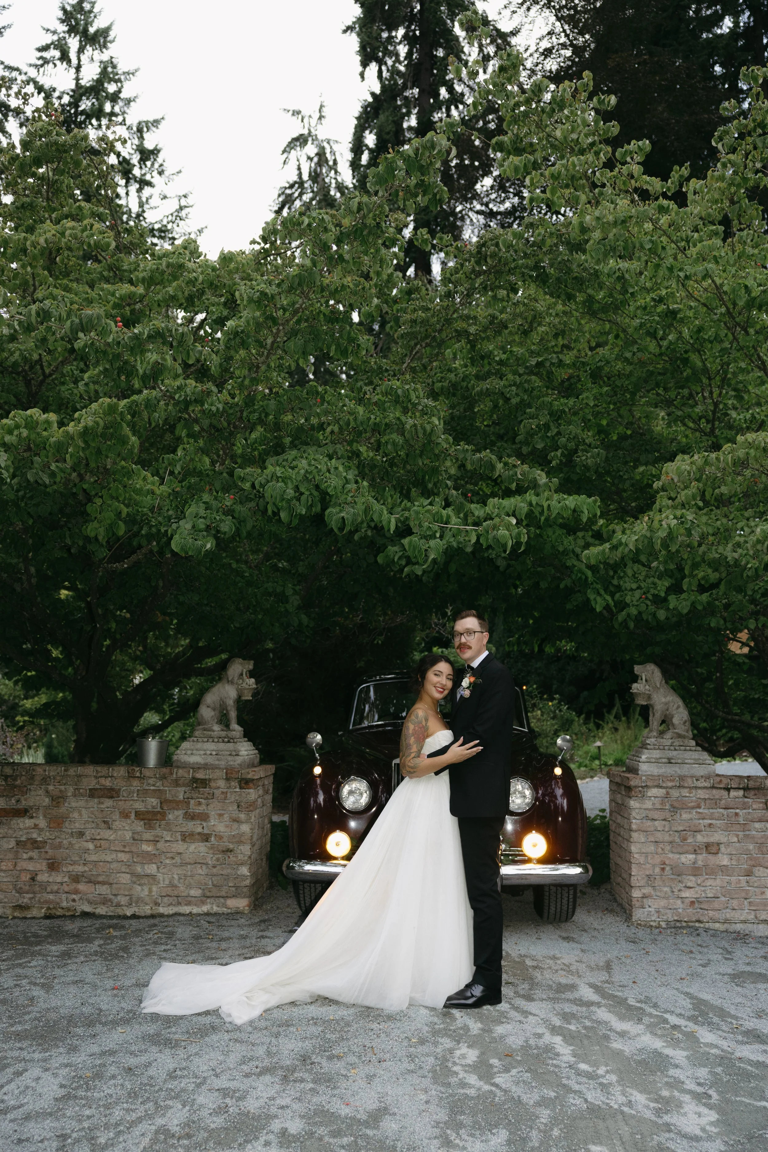 A bride and groom in wedding attire standing in front of a vintage car, with brick pillars topped with stone dog statues and green trees in the background.