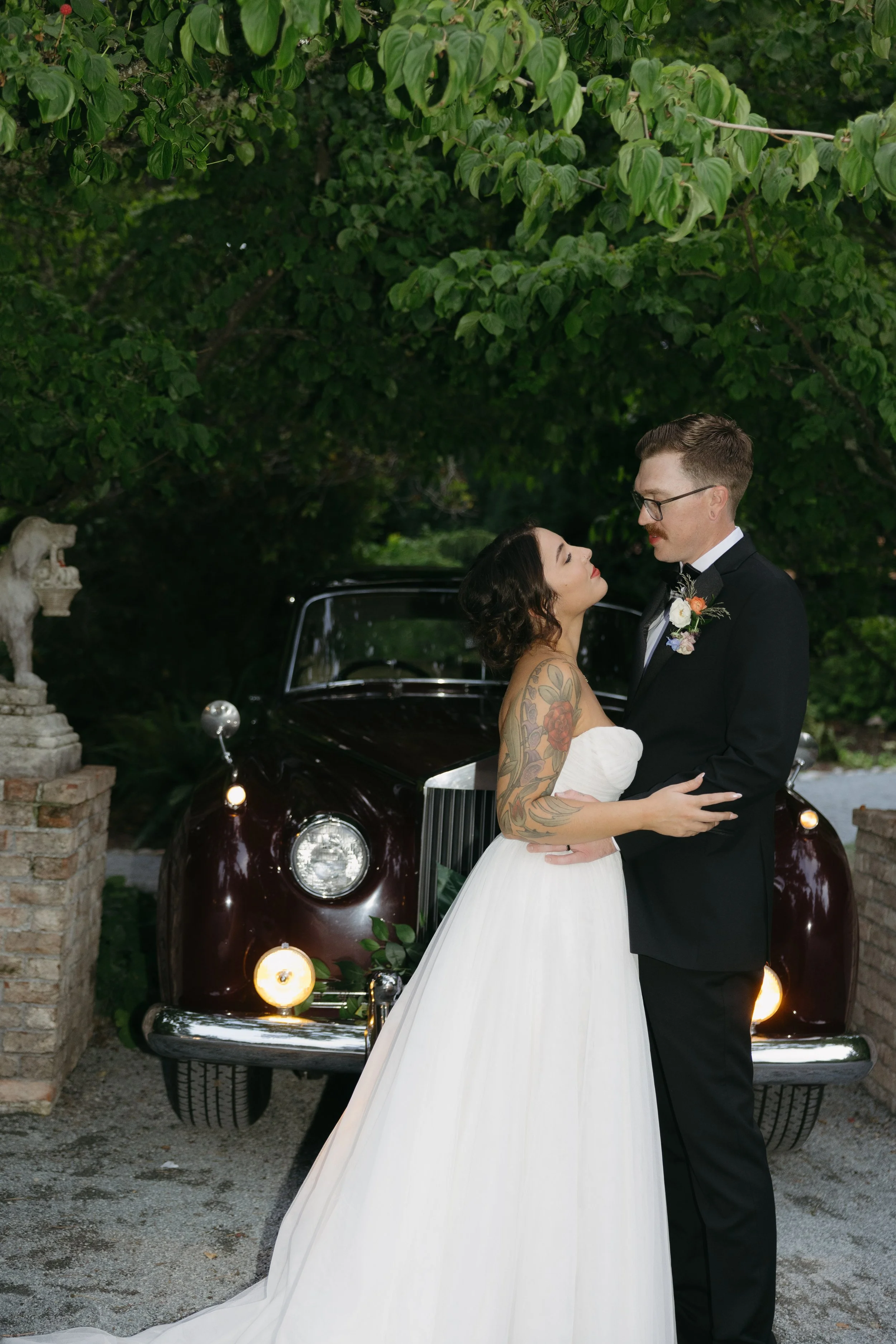 A bride and groom embracing outdoors near a vintage car, with green trees overhead.