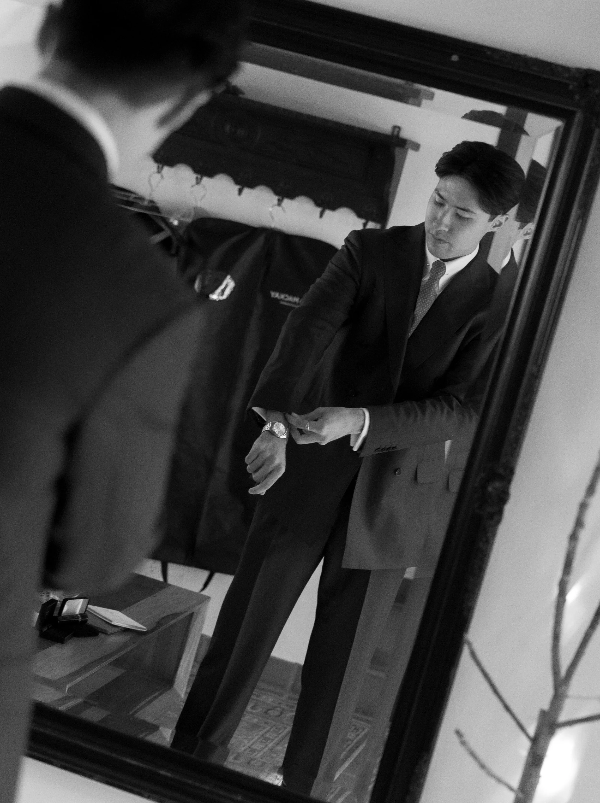 A man in a suit looking at his watch in a mirror, with another person out of focus in the foreground.