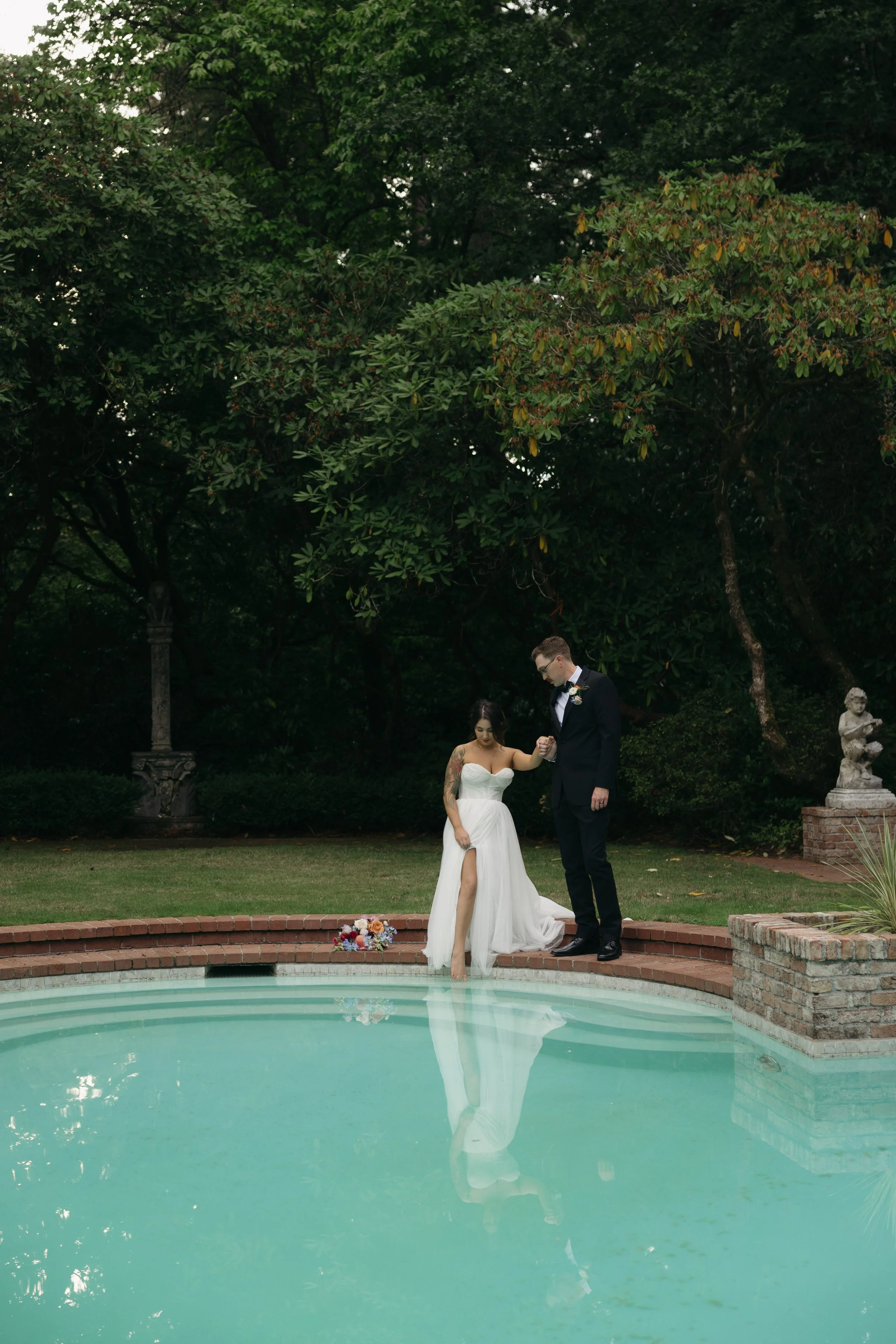 Bride and groom standing by a swimming pool, holding hands, outdoors in a lush garden, with a bouquet of flowers on the ground nearby at a European inspired Lakewold Gardens wedding in Seattle