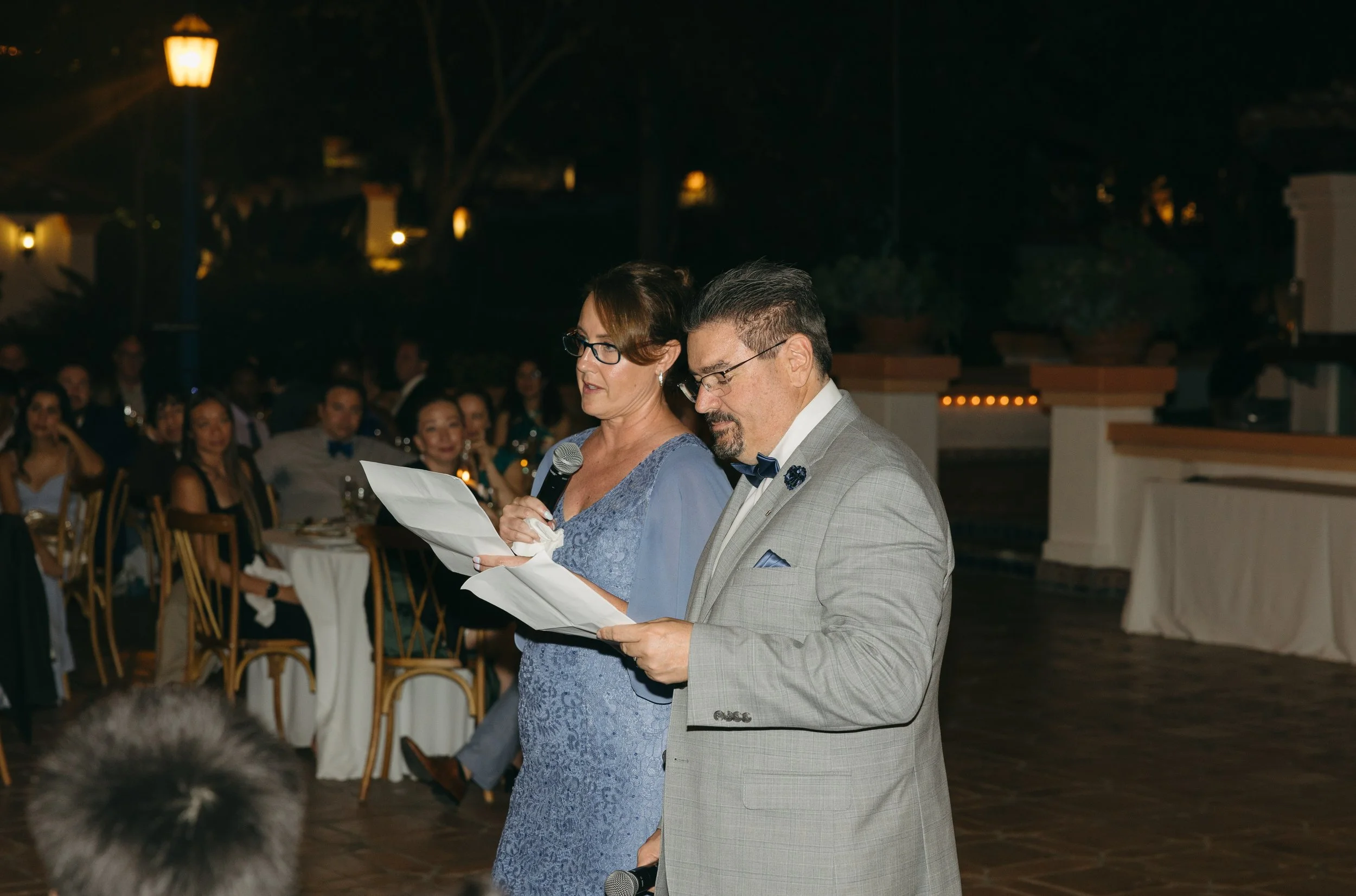 A woman and a man are reading from sheets of paper at an outdoor event during nighttime. The woman is holding a microphone and wearing a blue dress with lace details, glasses, and has short brown hair. The man is dressed in a light grey tuxedo with a