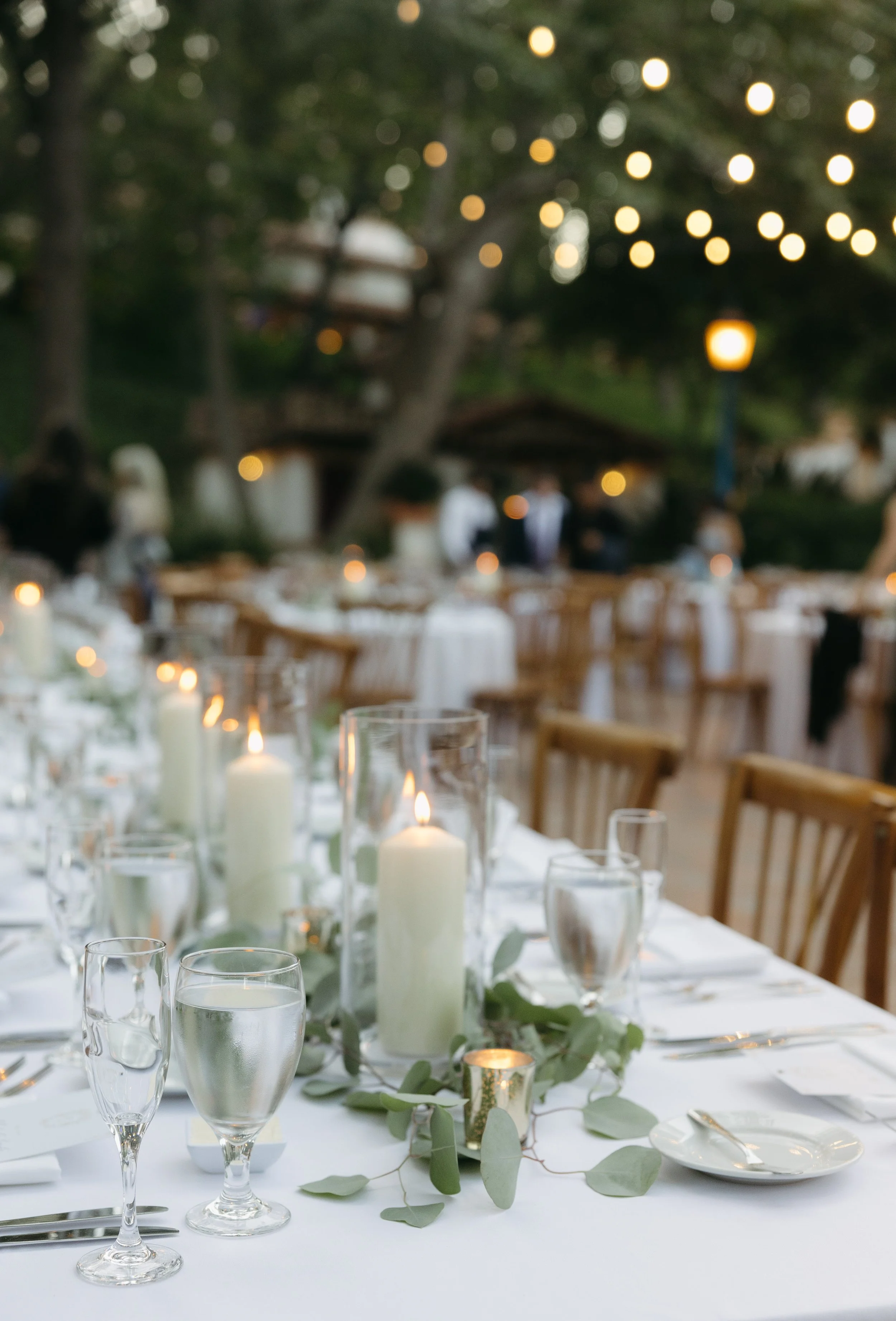 Wedding reception outdoor table with candles, greenery, glassware, and plates, set for an elegant event with blurred string lights and guests in the background at a Rancho Las Lomas Wedding in Los Angeles