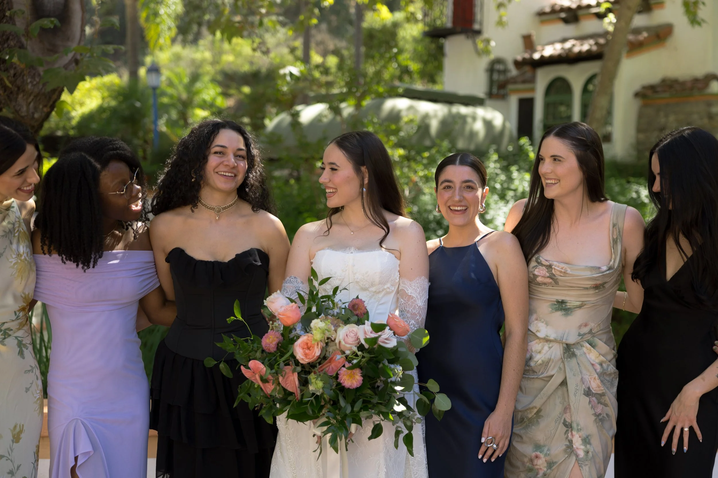 Group of bridesmaids dressed in formal attire, gathered outdoors during a wedding celebration, standing together with the bride holding a bouquet of flowers, all smiling and enjoying the moment.