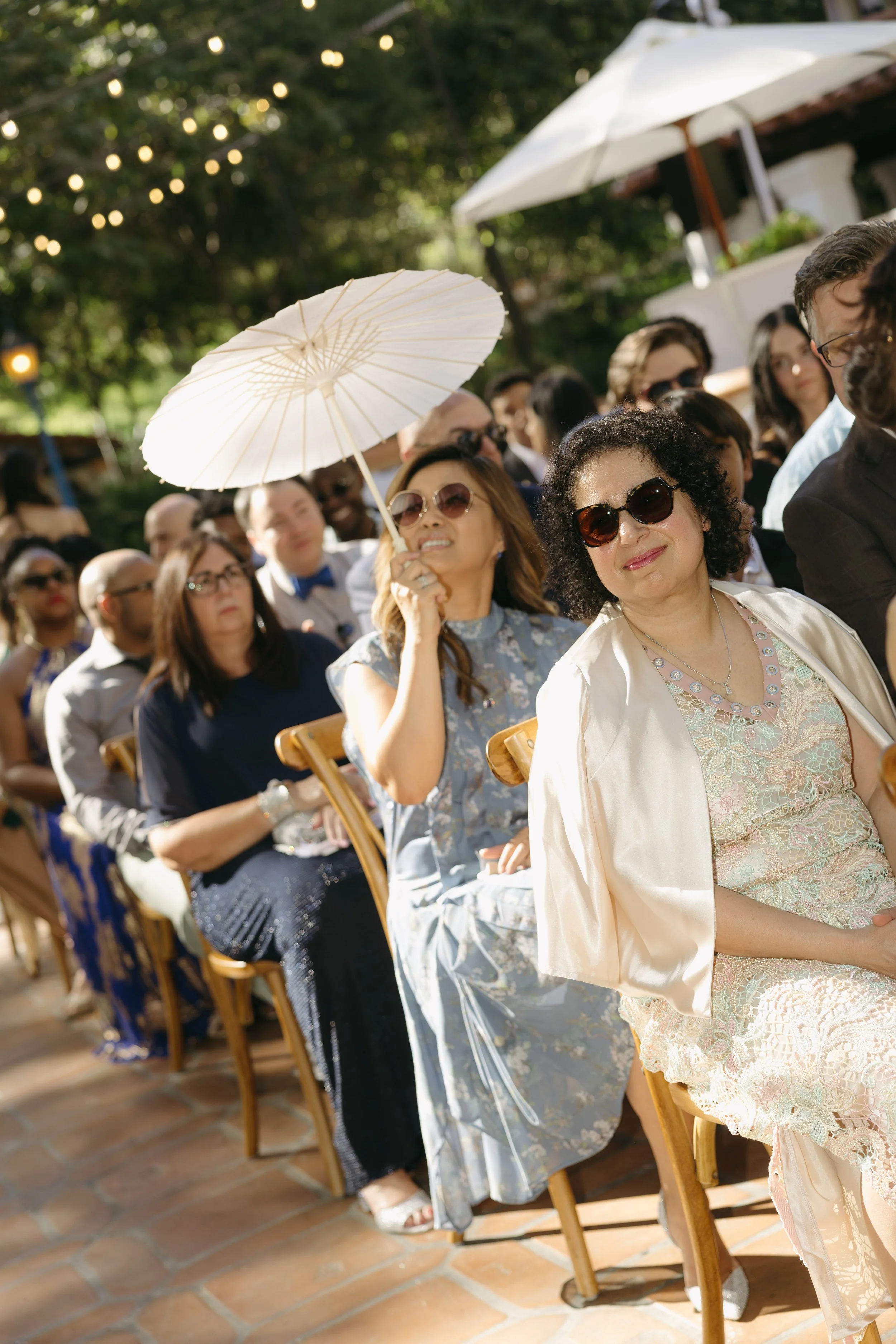 People seated at an outdoor wedding ceremony, some wearing sunglasses, during a sunny event. A woman in the foreground holds a parasol and smiles at a Rancho Las Lomas Wedding in Los Angeles