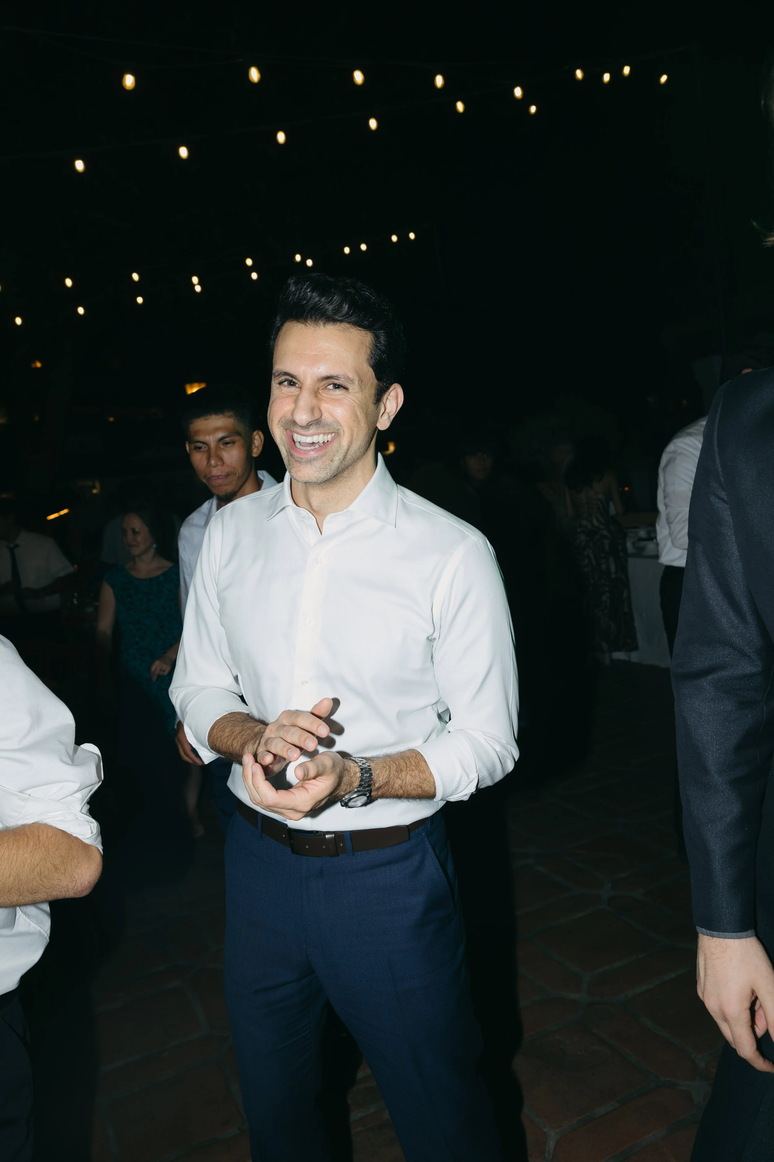 A man in a white shirt smiling and clapping at a social event in a dimly lit venue with string lights overhead.