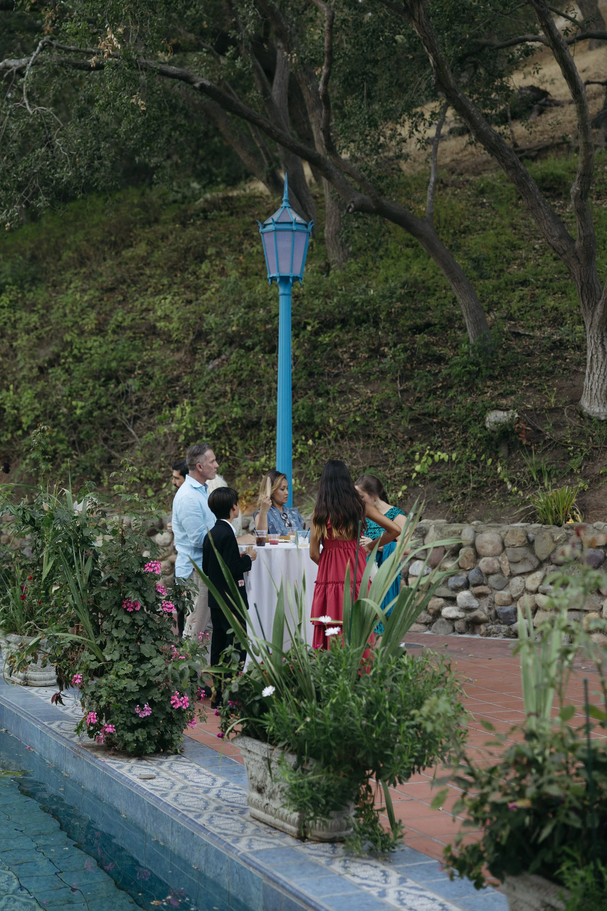 Outdoor cocktail hour with guests near a table with drinks, surrounded by plants and flowers, with a blue streetlamp and trees in the background at a Rancho Las Lomas Wedding in Los Angeles