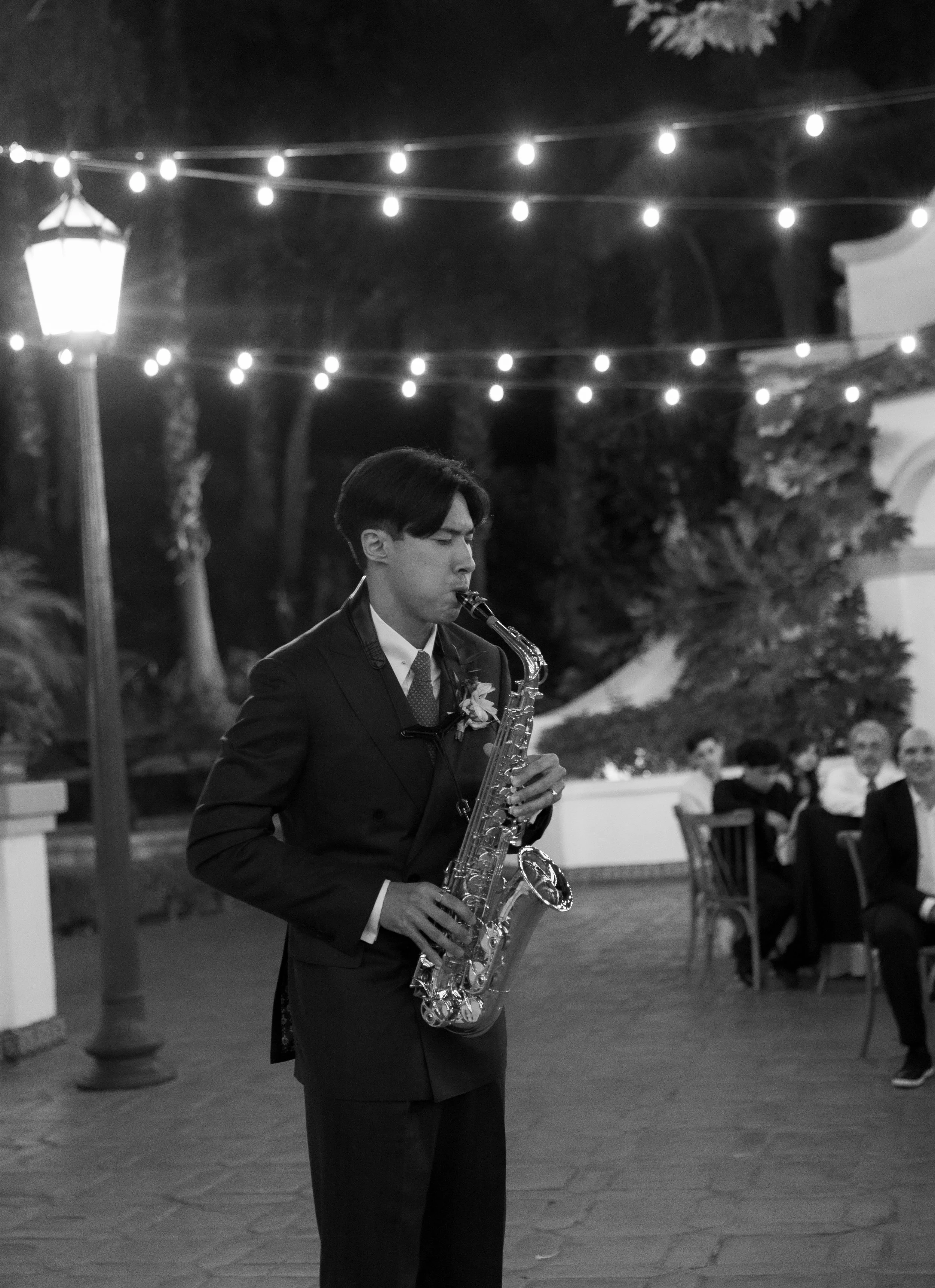 A man in a suit playing the saxophone at an outdoor evening event with string lights overhead and guests seated at tables in the background.