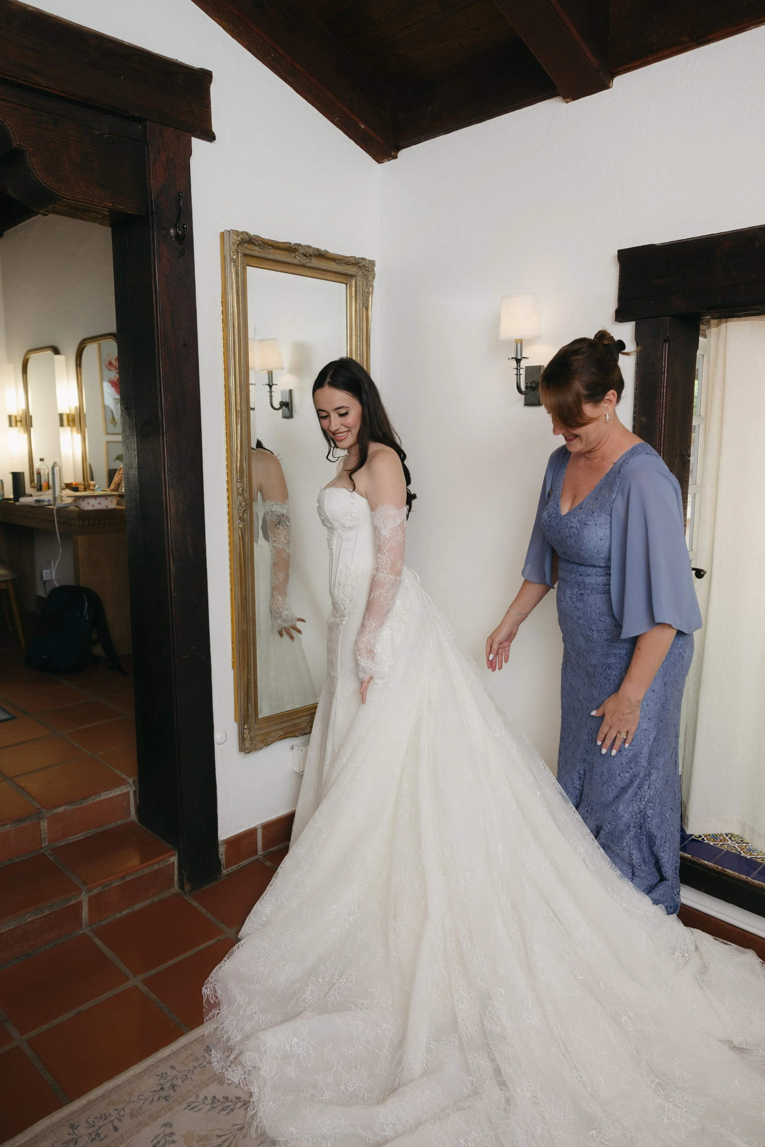 A bride in a white lace wedding gown adjusting her dress in front of a mirror, with a woman in a blue dress assisting her, in a warmly lit room with dark wood beams and terracotta tile flooring.