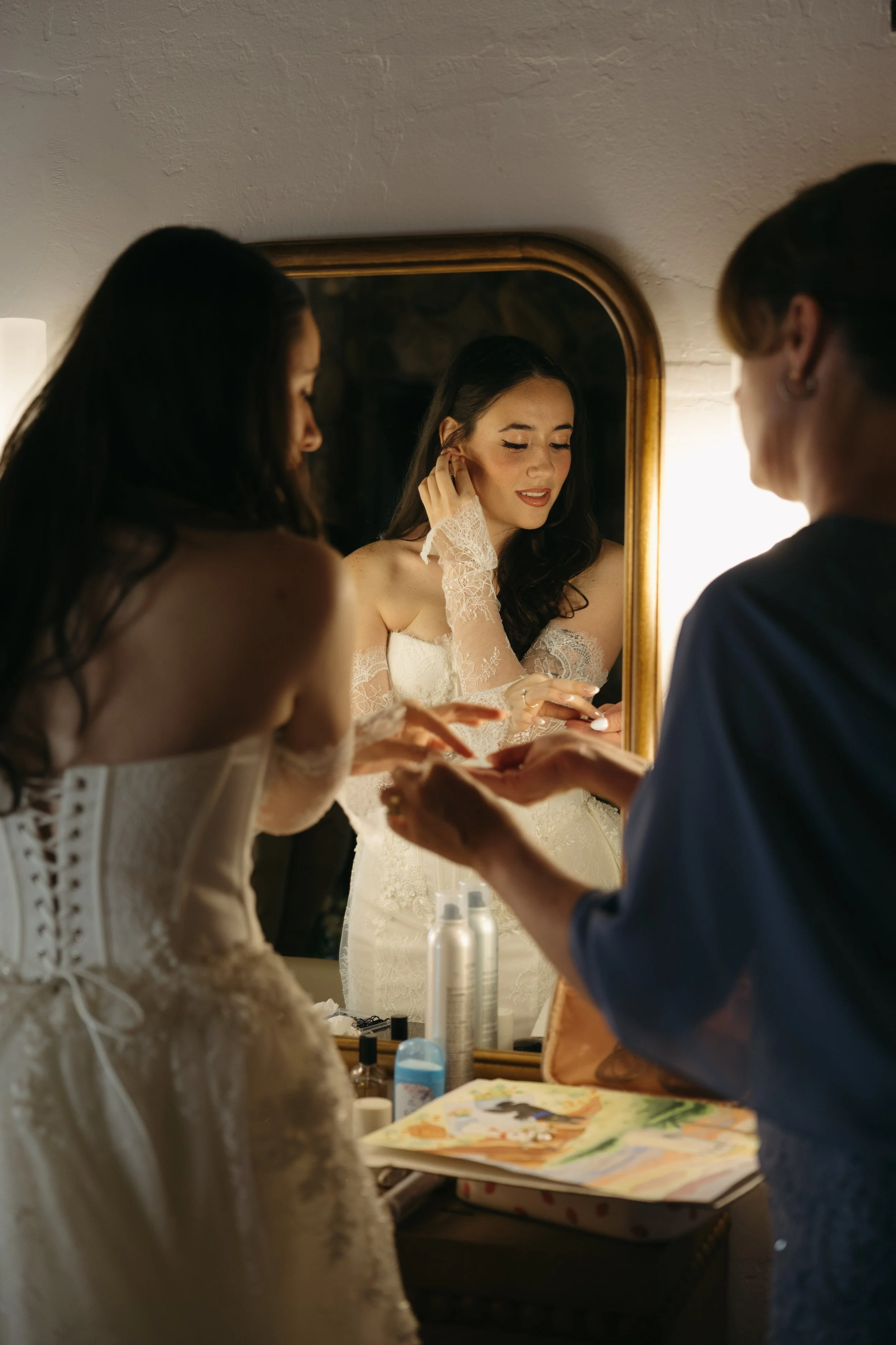A bride in a wedding dress gets ready with help from two women in front of a mirror, applying makeup and preparing her for a Rancho Las Lomas Wedding in Los Angeles