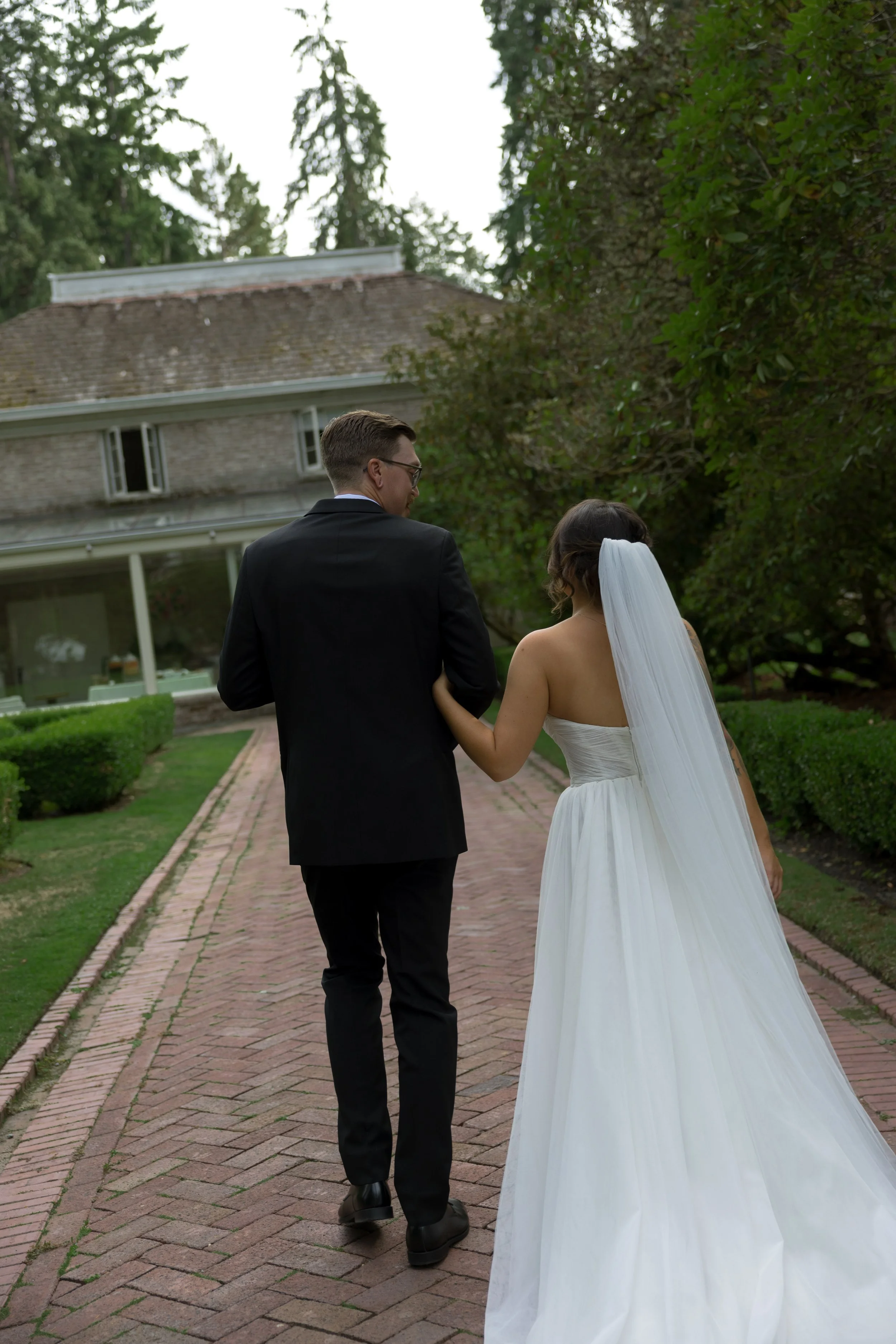 A bride and groom walking arm in arm down a brick pathway surrounded by greenery and trees, outside a house.