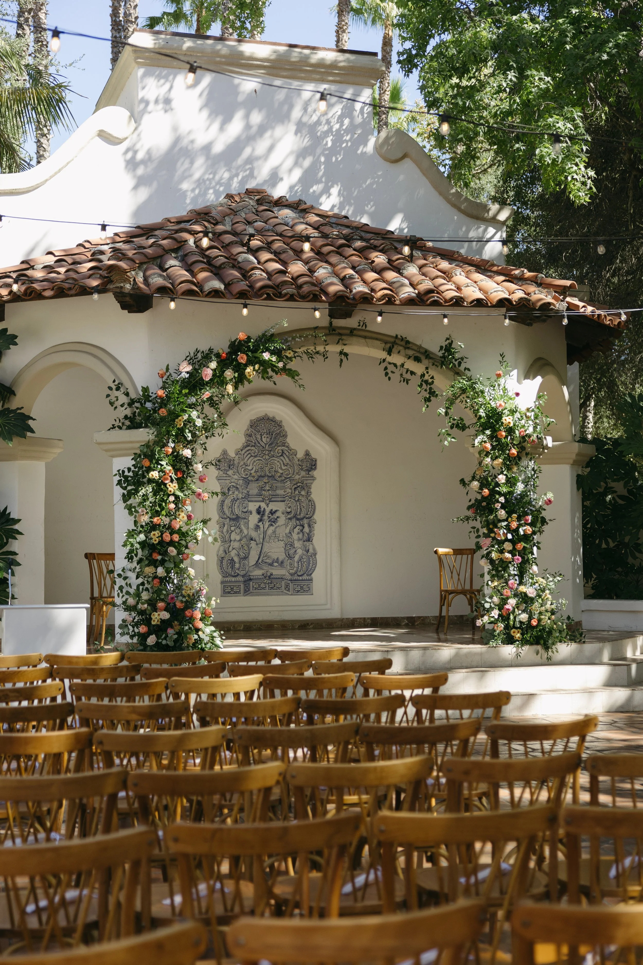 Outdoor wedding ceremony setup with a small stage decorated with floral arrangements and chairs arranged in front of it, under string lights and trees at a Rancho Las Lomas Wedding in Los Angeles