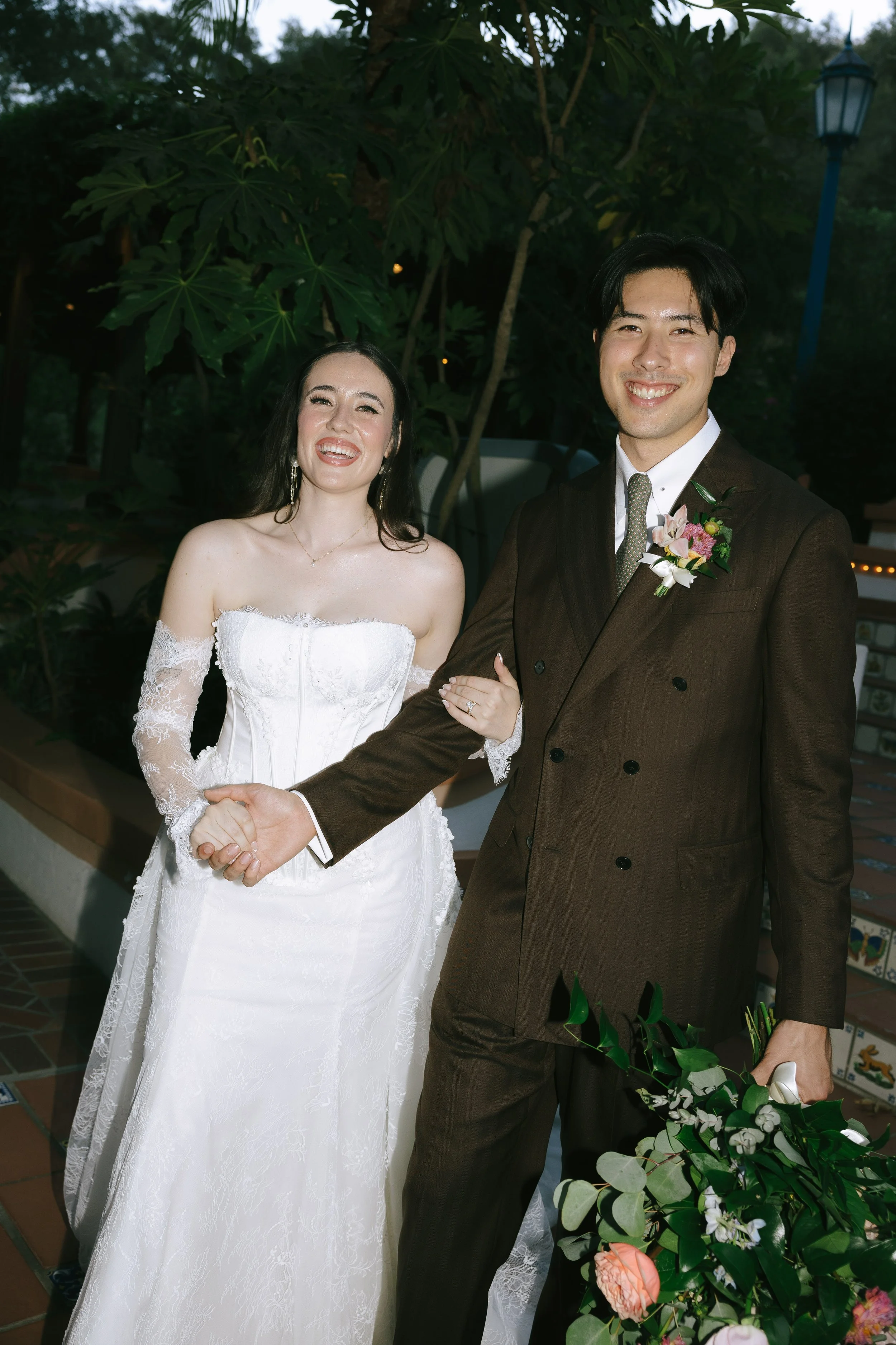 A smiling bride and groom holding hands, standing outdoors with greenery and a blue lamppost in the background.