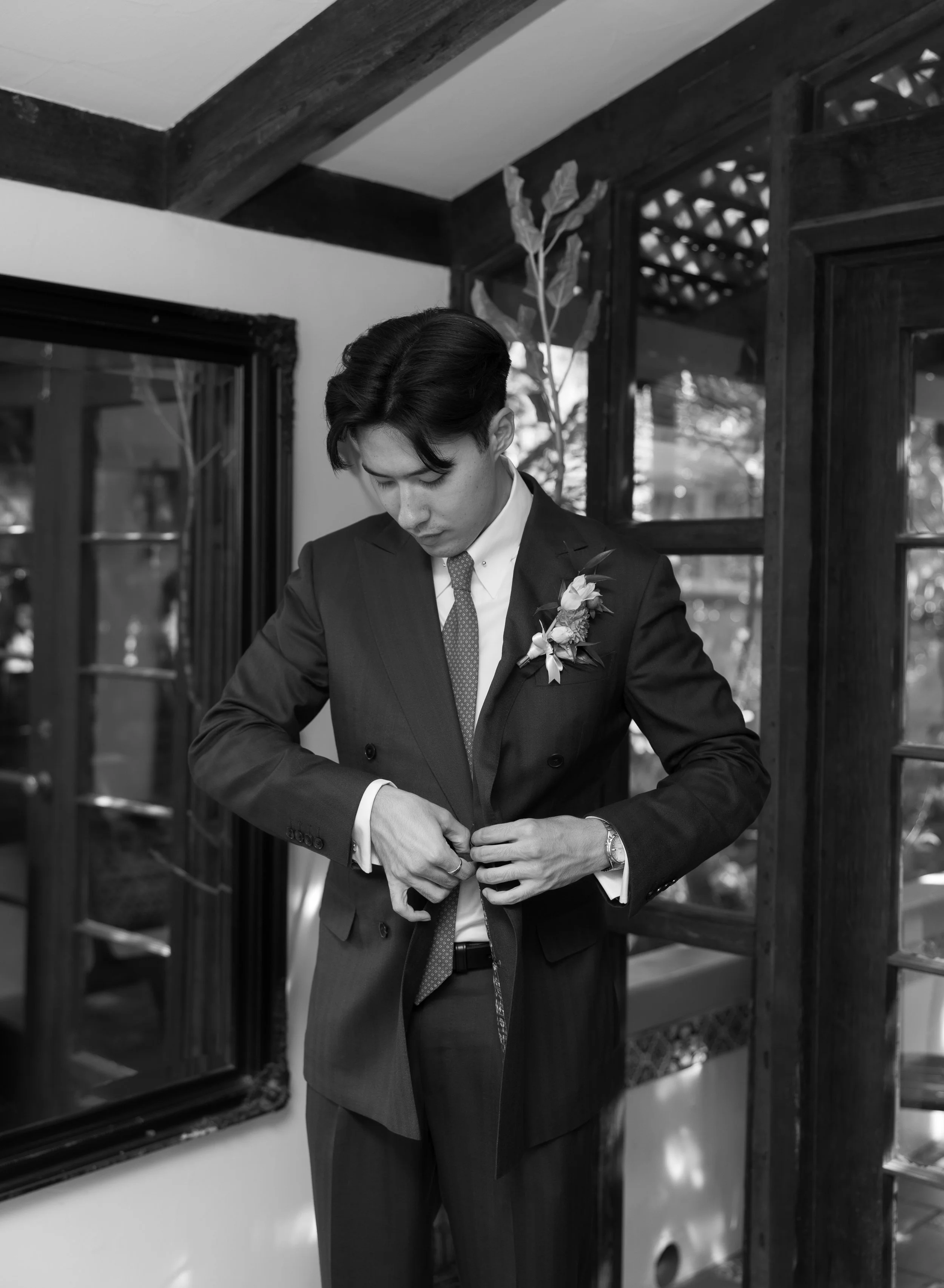 A man in a suit adjusting his jacket inside a room with wooden window frames and a decorative plant.