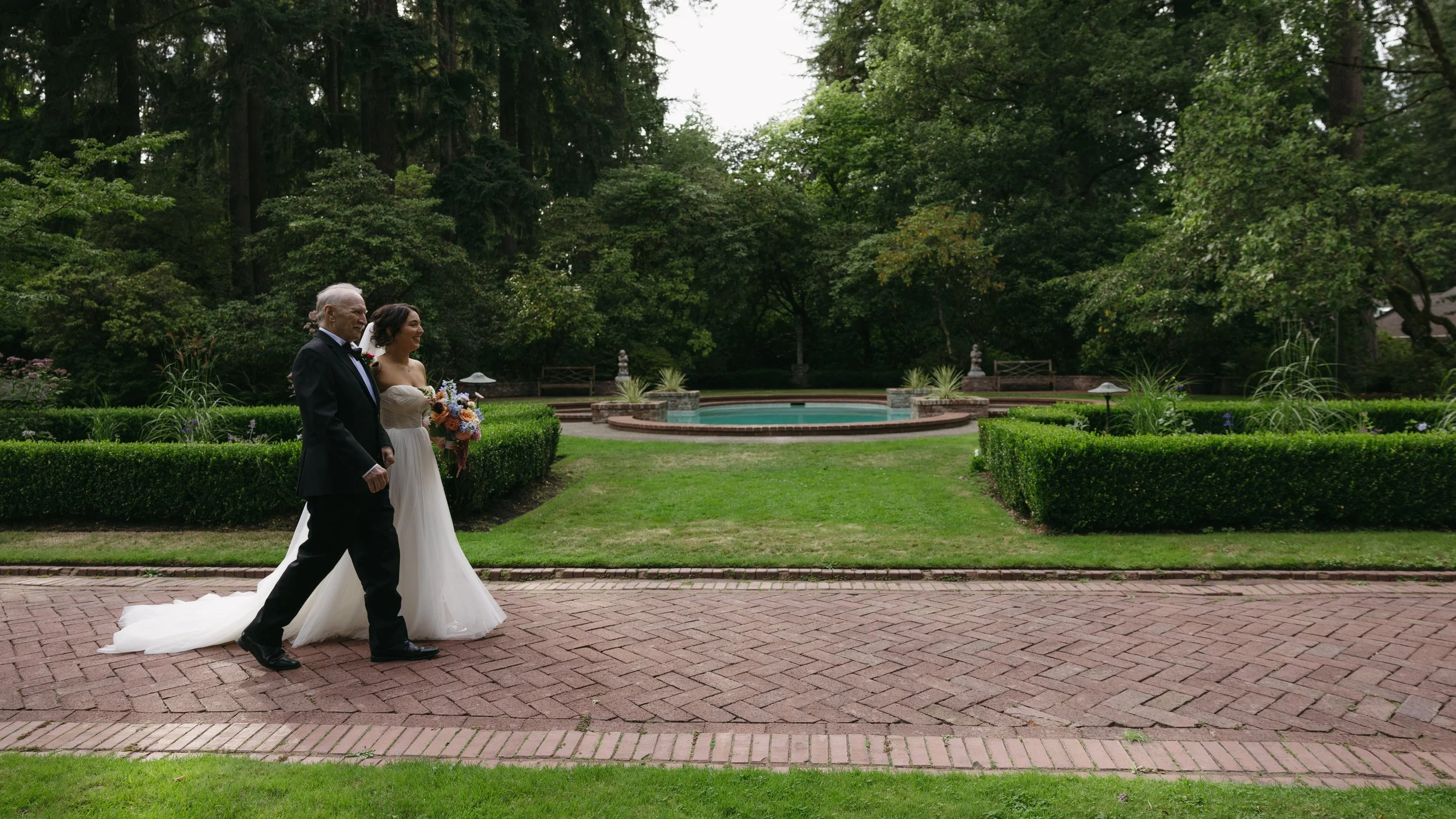 Bride in a wedding dress walking with her father in a garden with lush greenery, a pond, and flower arrangements at a European inspired Lakewold Gardens wedding in Seattle