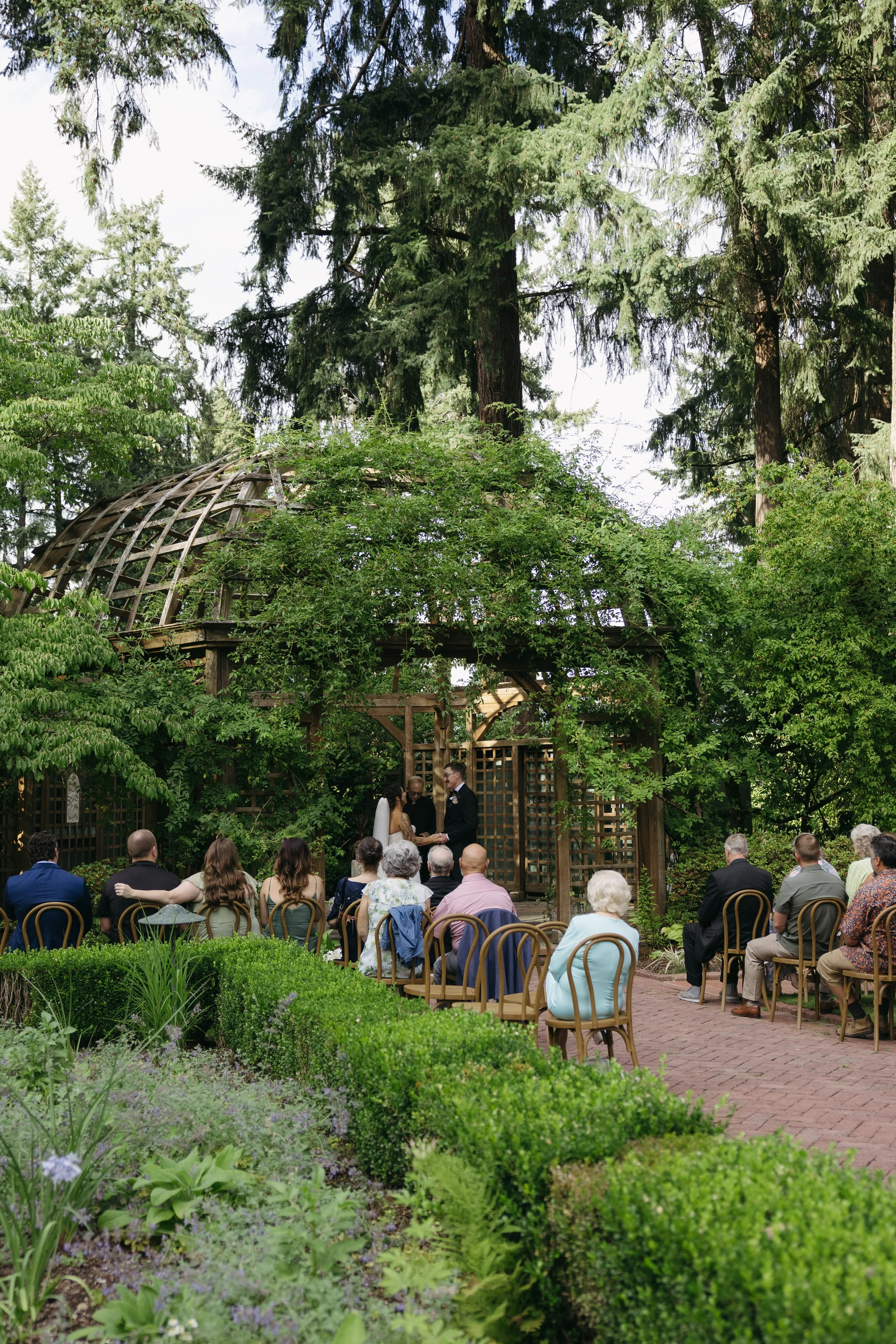A wedding ceremony taking place outdoors under a wooded structure with tall trees, as guests sit in chairs watching the couple exchange vows at a European inspired Lakewold Gardens wedding in Seattle