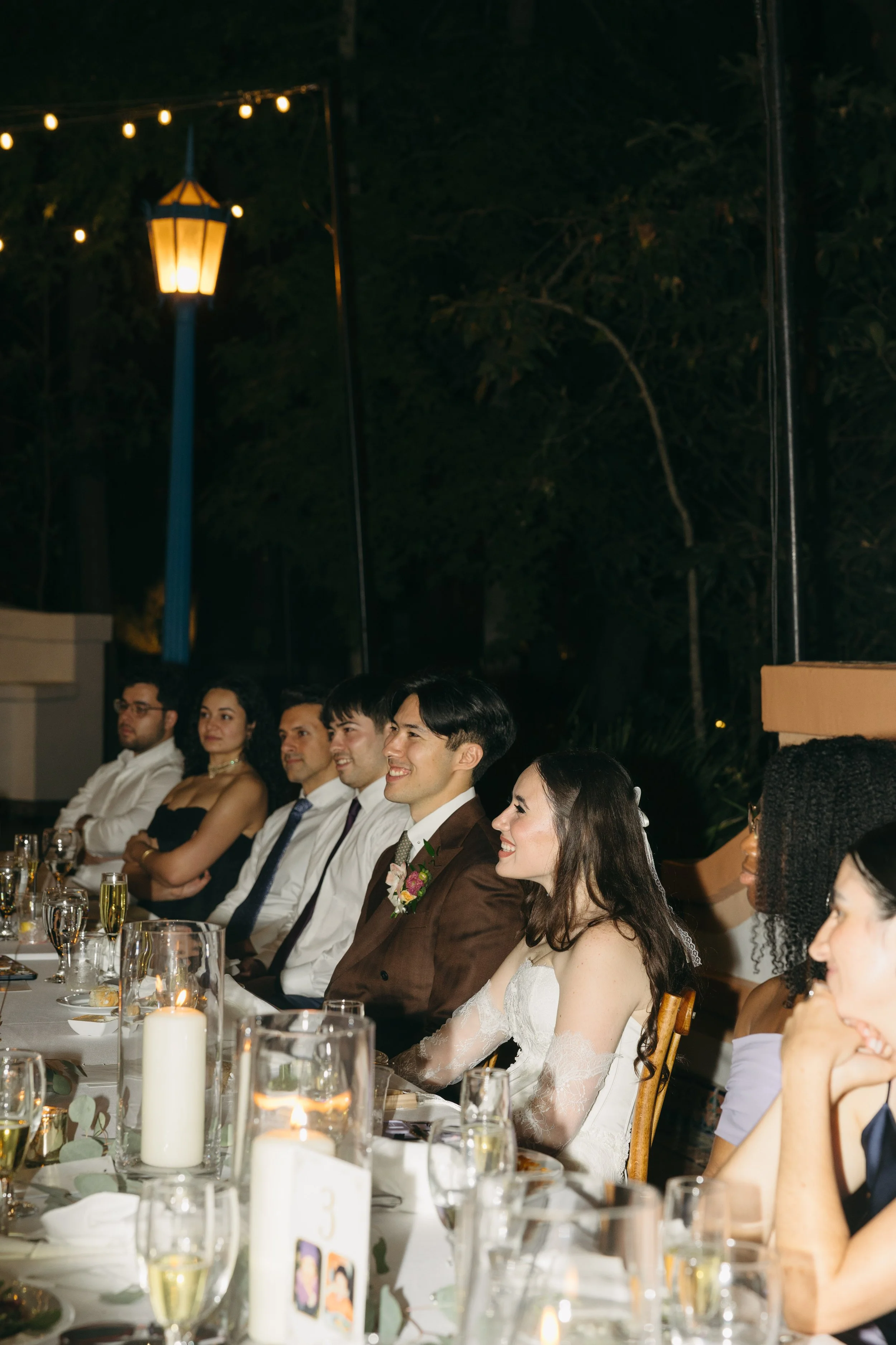 People sitting at a long table during an outdoor evening event, with string lights and a lamp in the background