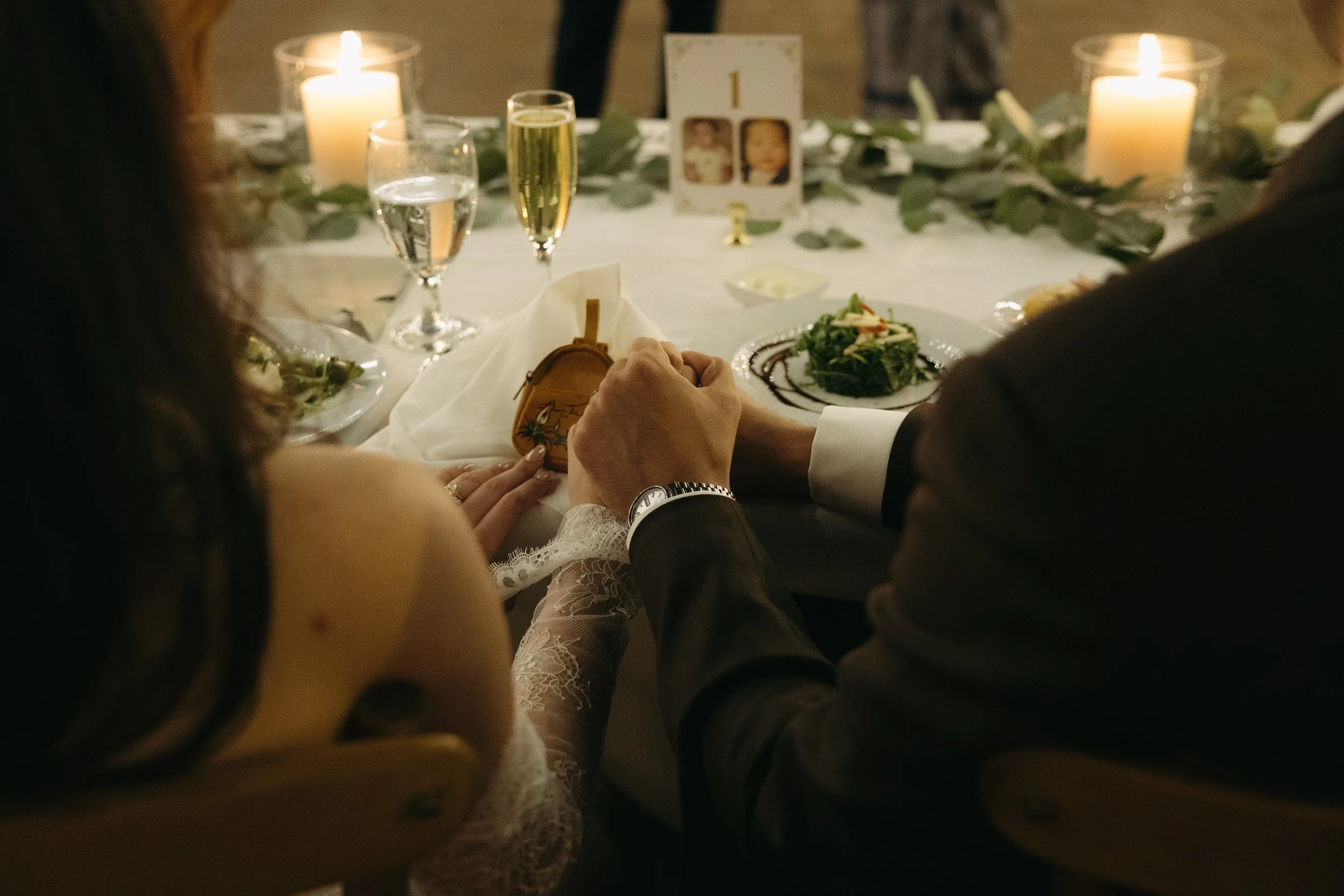 A couple holding hands at a formal dinner table with candles, wine glasses, and a salad, during a romantic event.