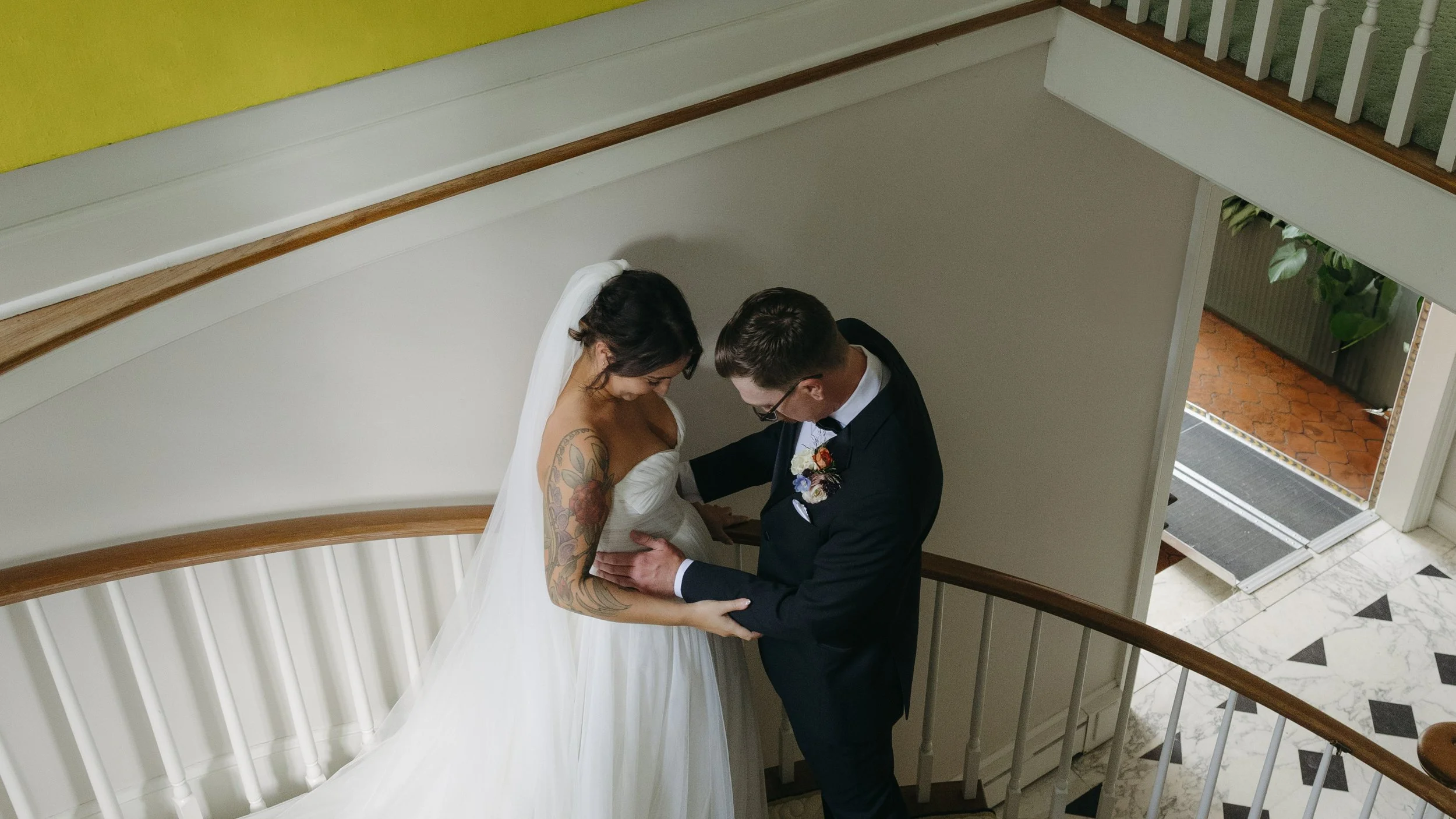 A bride and groom sharing a moment on a staircase inside a house, with the bride wearing a white wedding gown and with tattoos on her arm, and the groom in a dark suit and glasses.