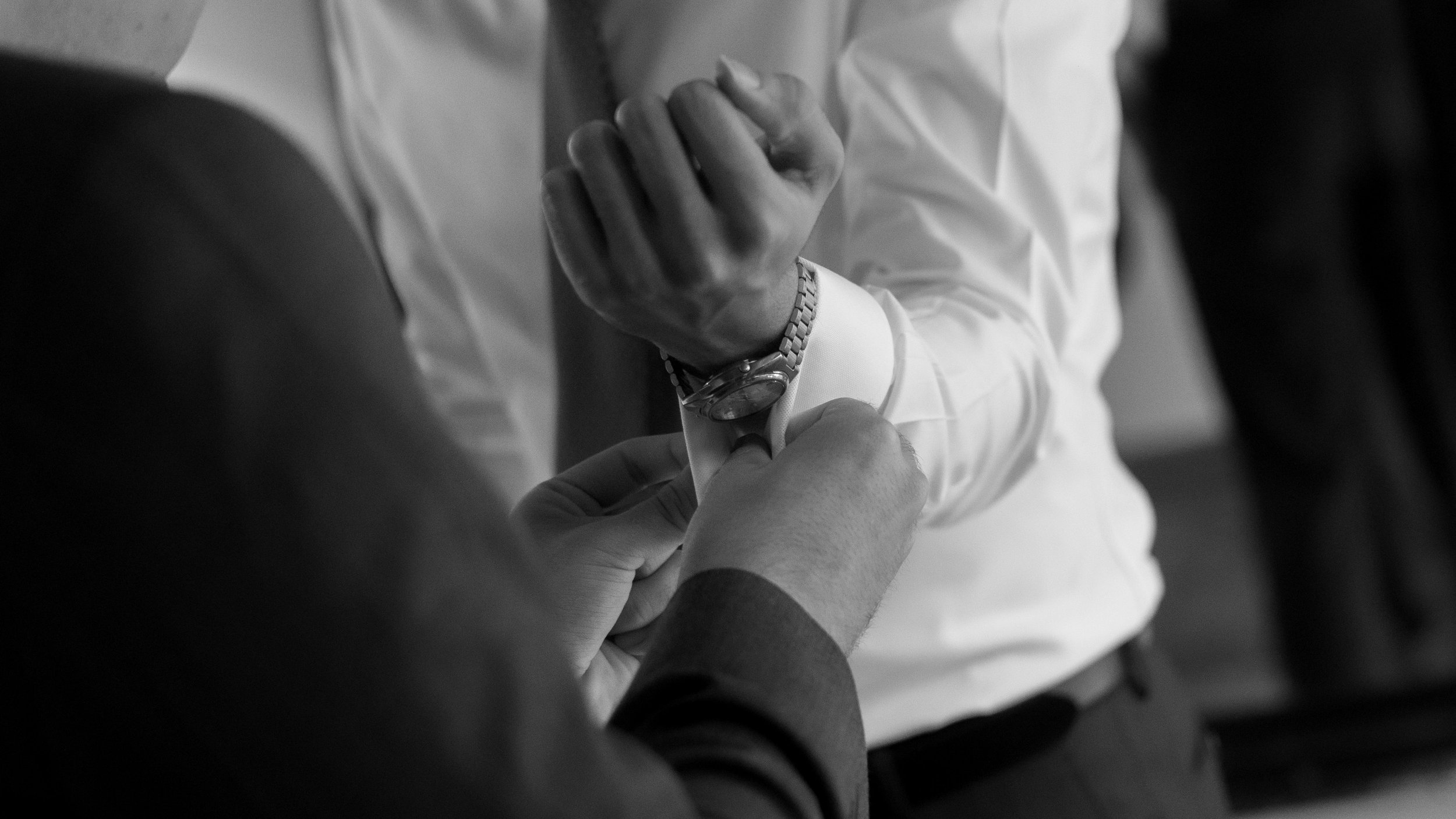 A person adjusting the cuff of another person's shirt sleeve, with the focus on their hands and wristwatch in black and white.
