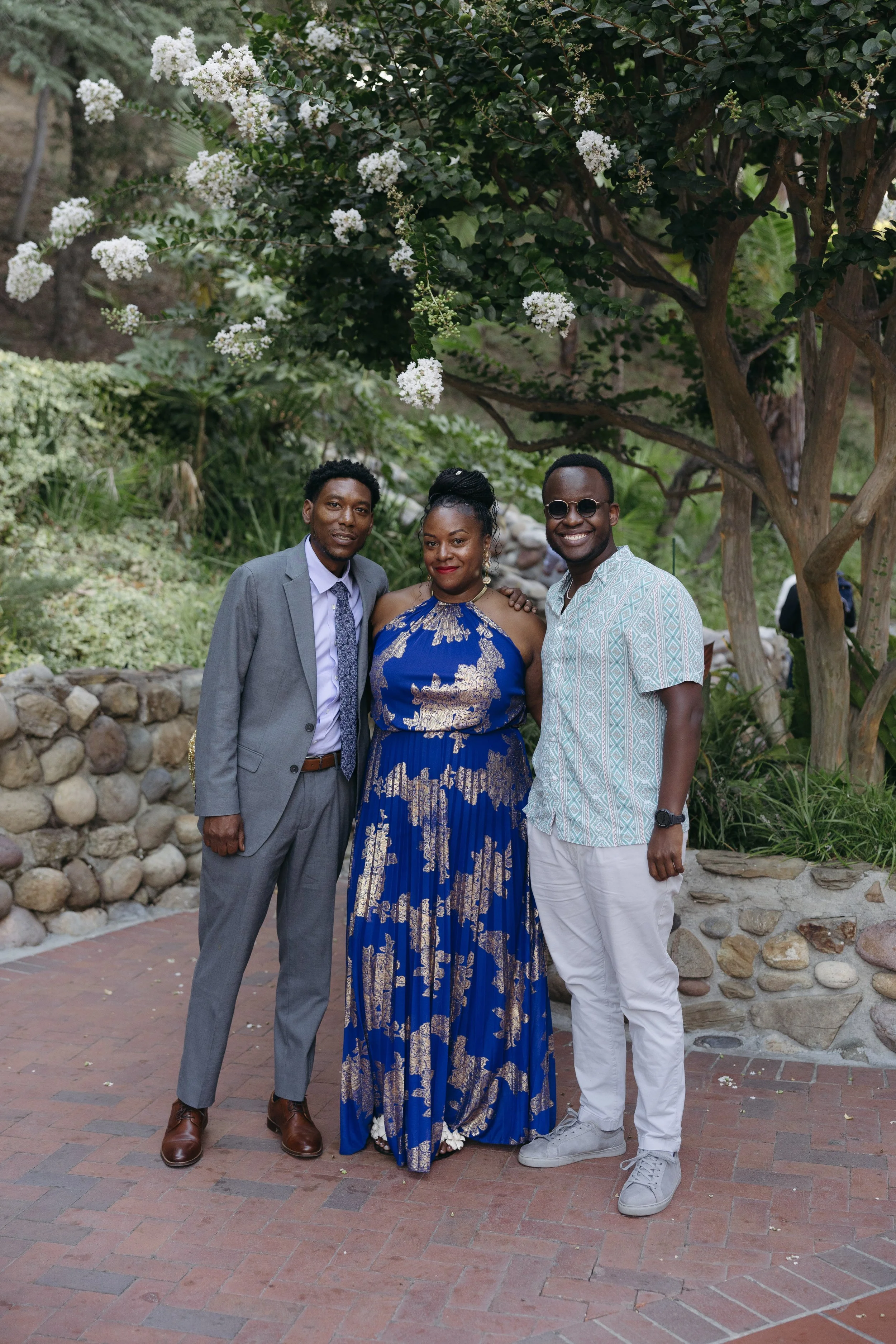 Three people standing outdoors under a flowering tree, dressed in semi-formal attire, smiling.
