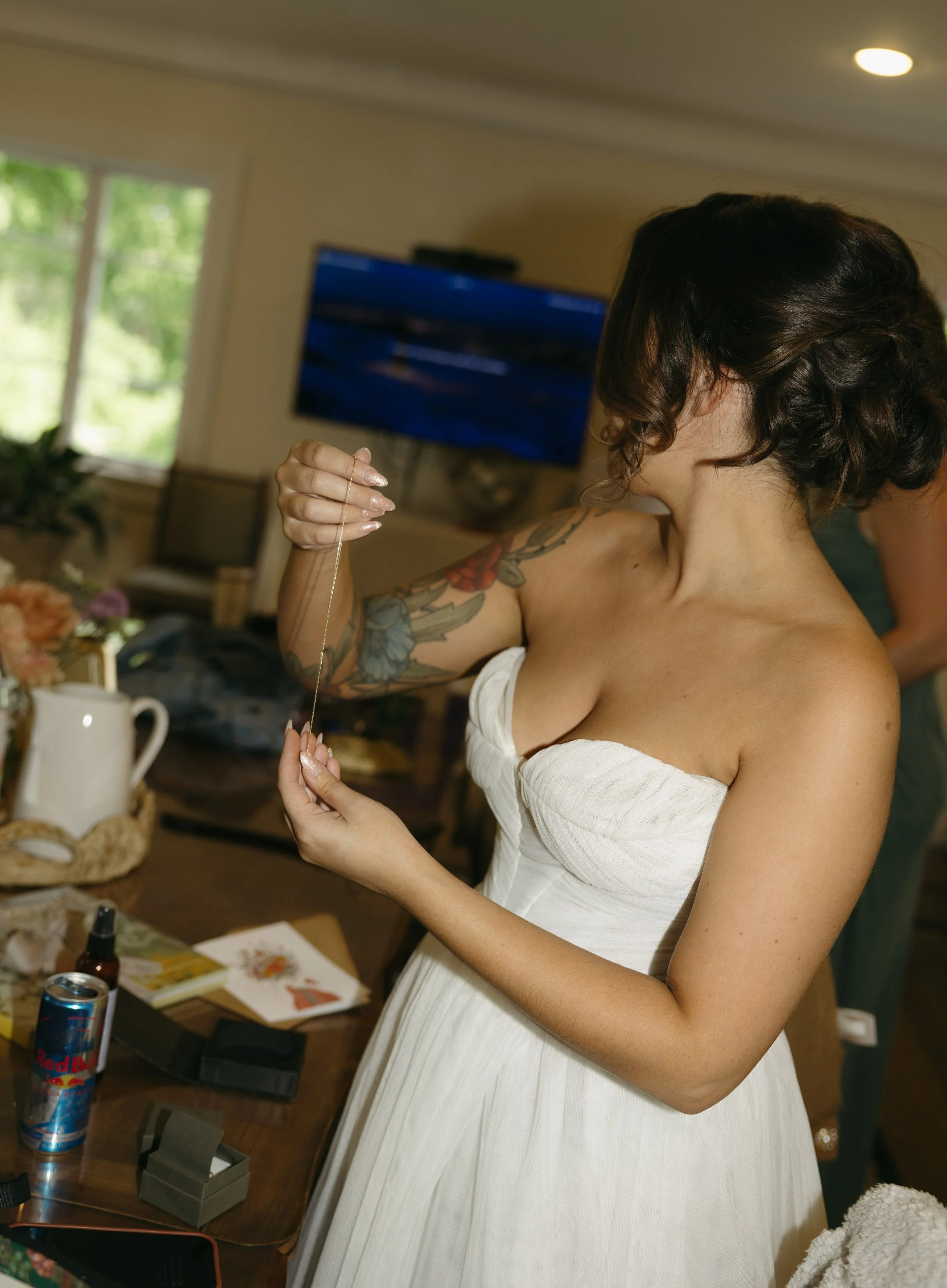 Woman in white strapless dress holding a necklace in a room with a TV, window, and various items on a table.