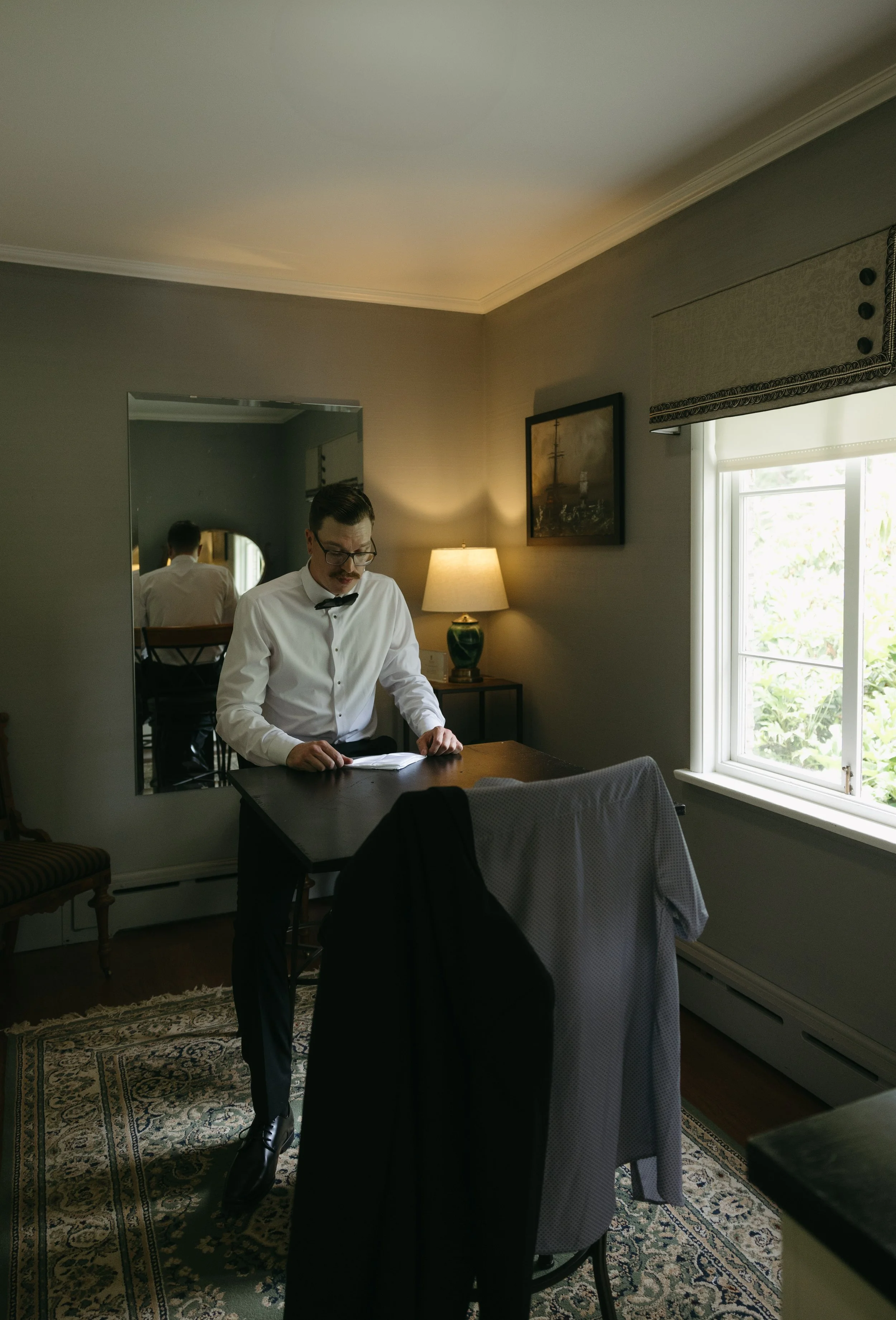 A man dressed in formal attire, including a white shirt and bow tie, standing at a table reading a book inside a well-lit room with a window, a lamp, a mirror, and a framed picture on the wall.