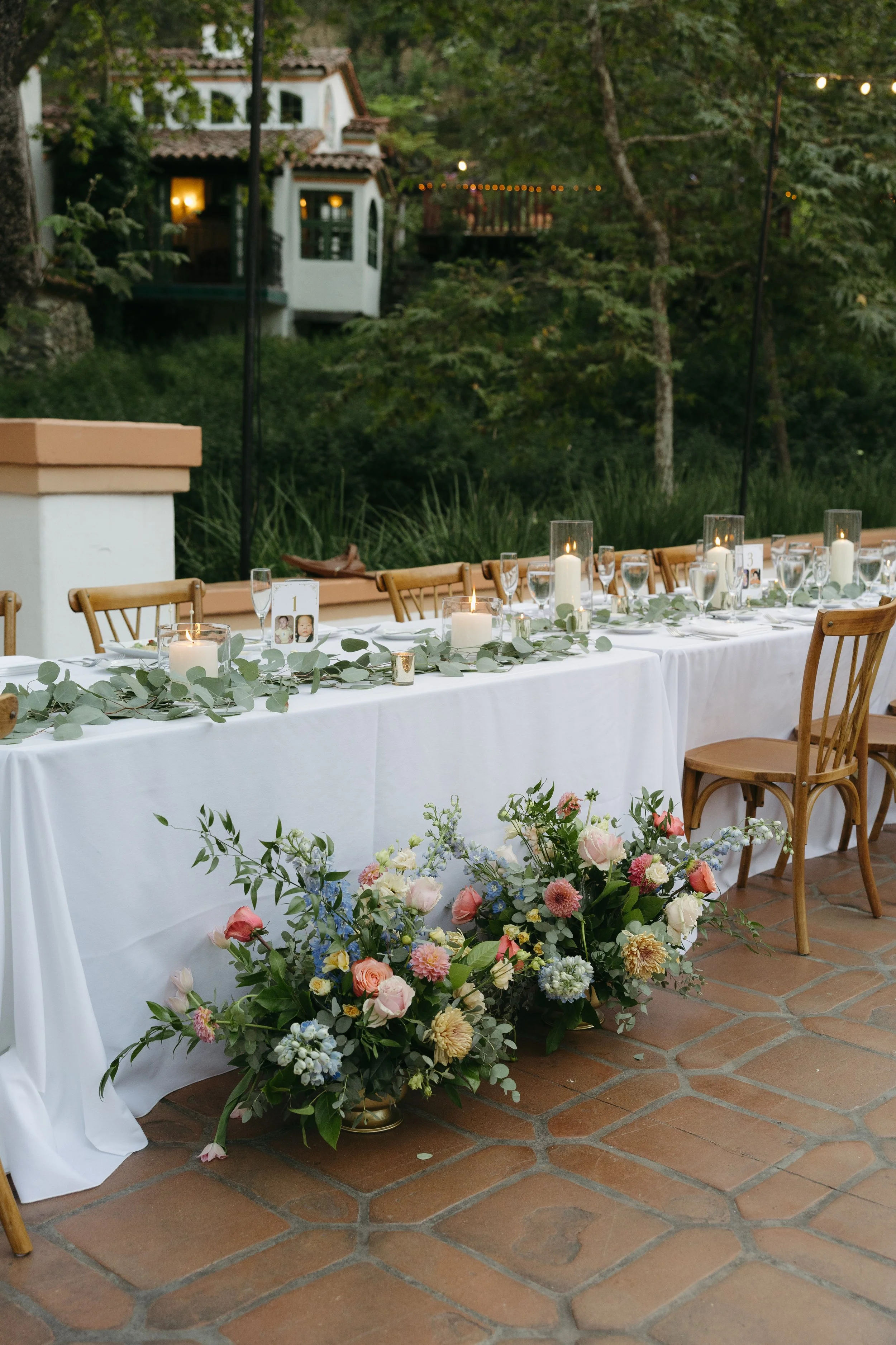 Wedding reception table decorated with white tablecloth, candles, glassware, and greenery, with a large floral arrangement at the end in an outdoor setting surrounded by trees and greenery at a Rancho Las Lomas Wedding in Los Angeles