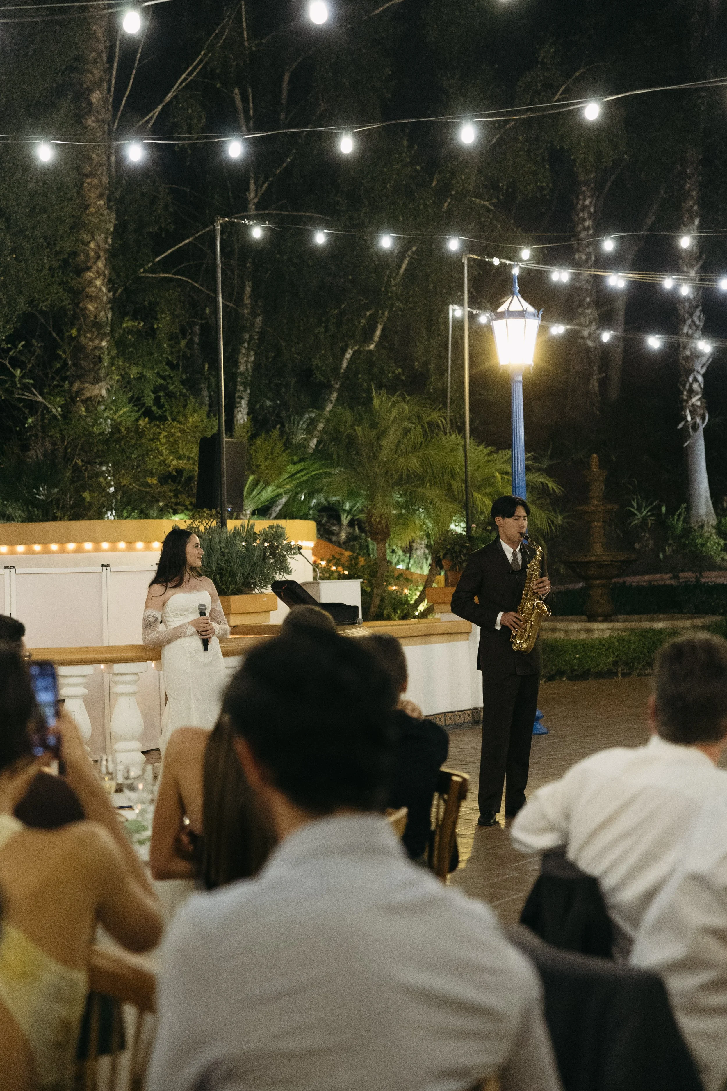 A music performance at an outdoor wedding reception during the evening, with a female singer in a white dress holding a microphone and a male saxophonist in a black suit playing saxophone, amidst string lights and a garden setting.