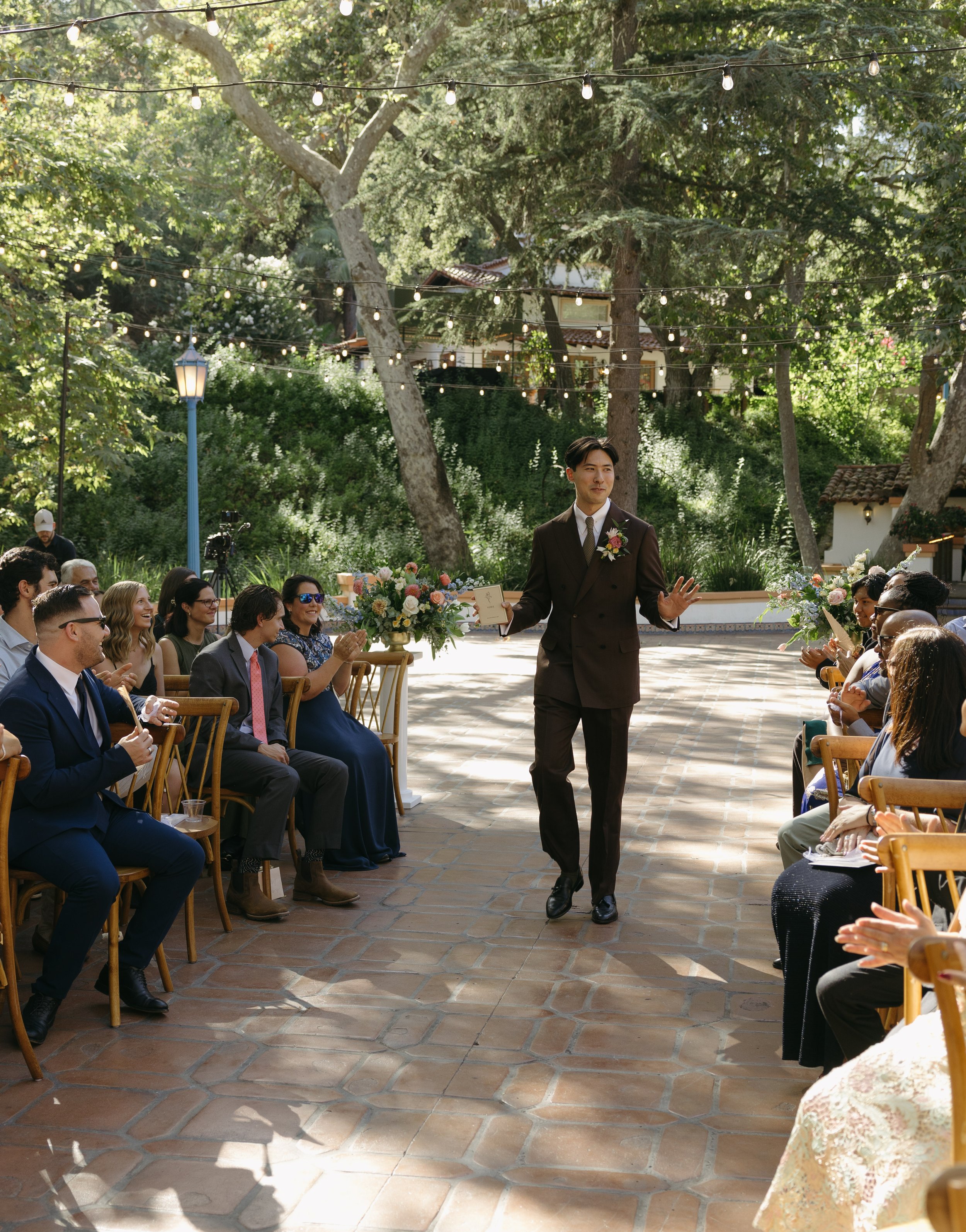 A wedding ceremony outdoors with a groom walking down the aisle, surrounded by seated guests clapping and smiling, under string lights hanging from trees at a Rancho Las Lomas Wedding in Los Angeles