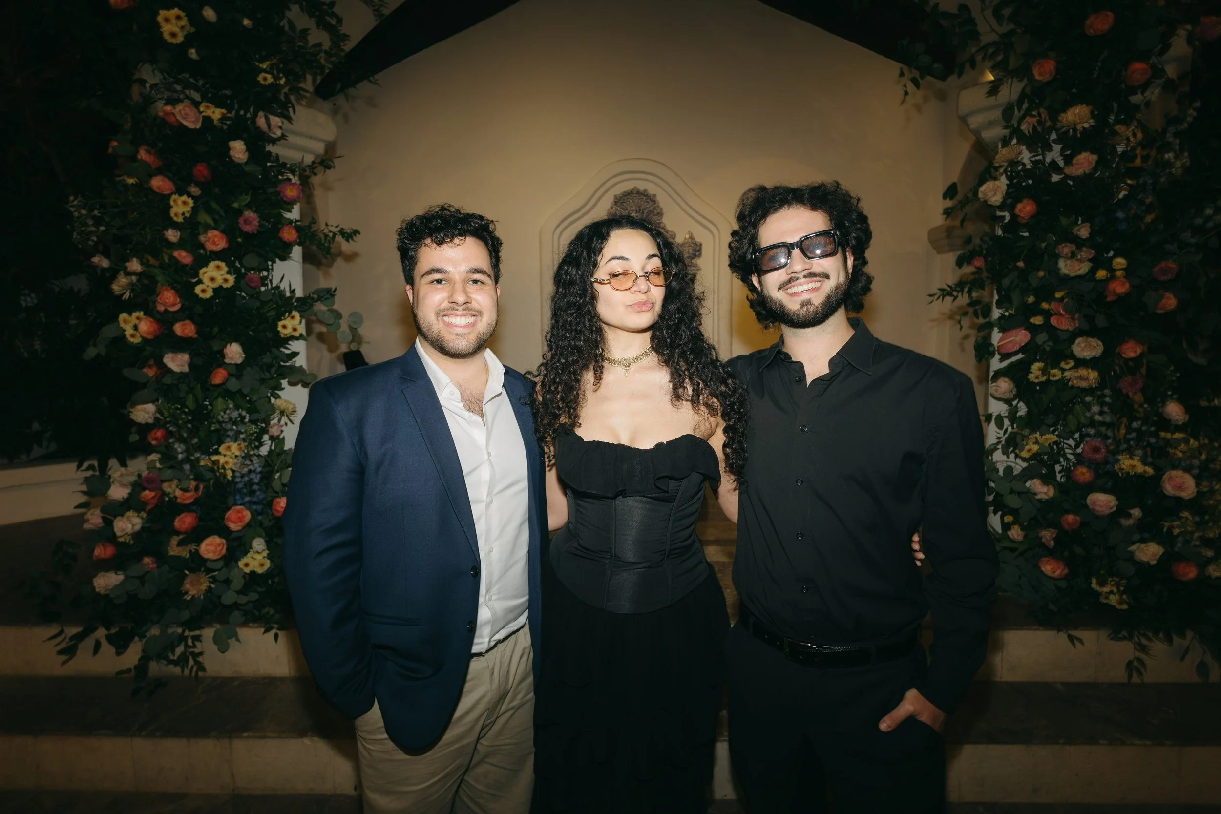 Three young adults, two men and one woman, standing together at a formal event, smiling, with a floral arch behind them.