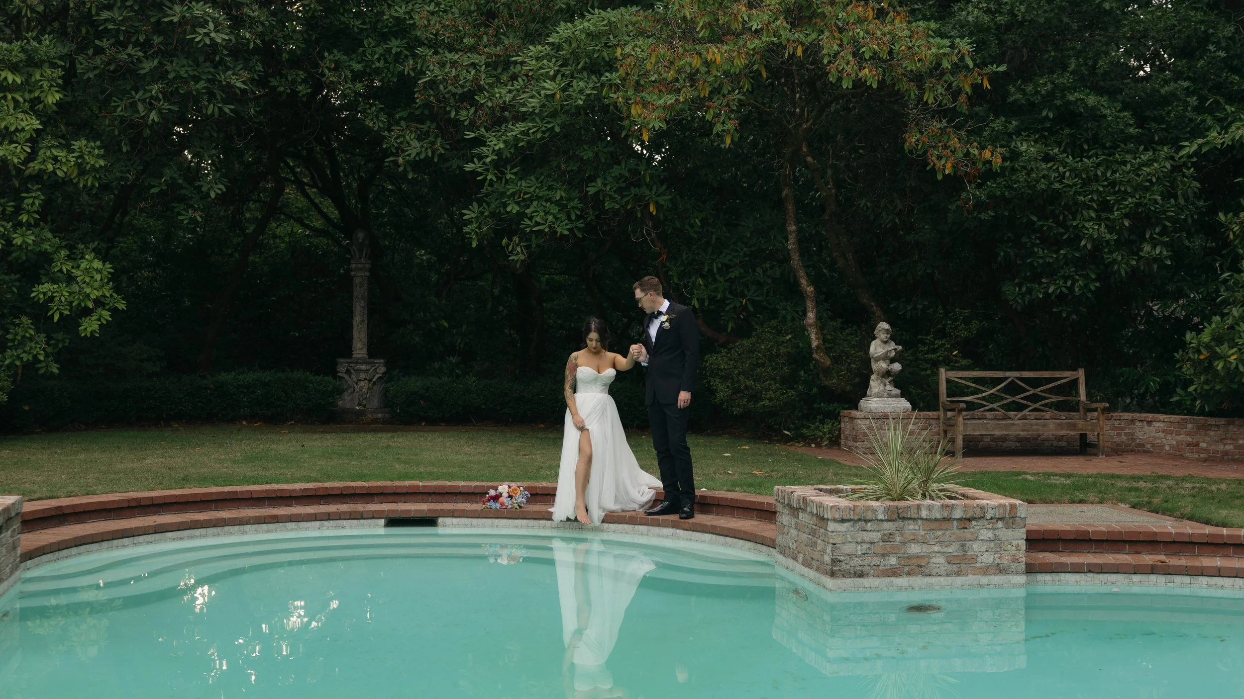 Bride and groom standing on poolside, holding hands, with wedding bouquet on ground, surrounded by lush green trees and garden decorations at a European inspired Lakewold Gardens wedding in Seattle