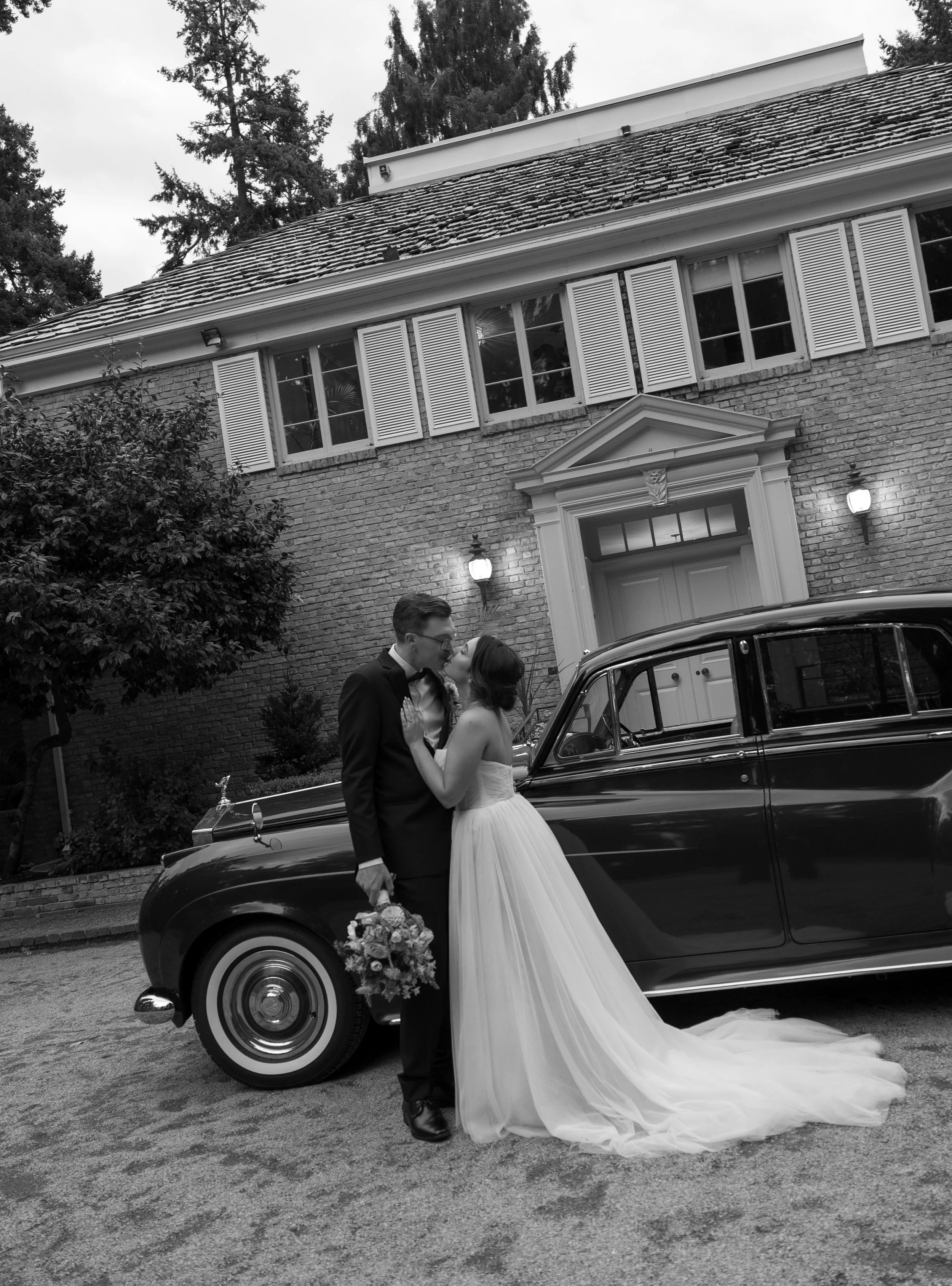 A bride and groom kissing in front of a vintage black car outside a brick house with white shutters. The bride is wearing a strapless wedding dress with a long train, and the groom is in a tuxedo holding a bouquet of flowers.