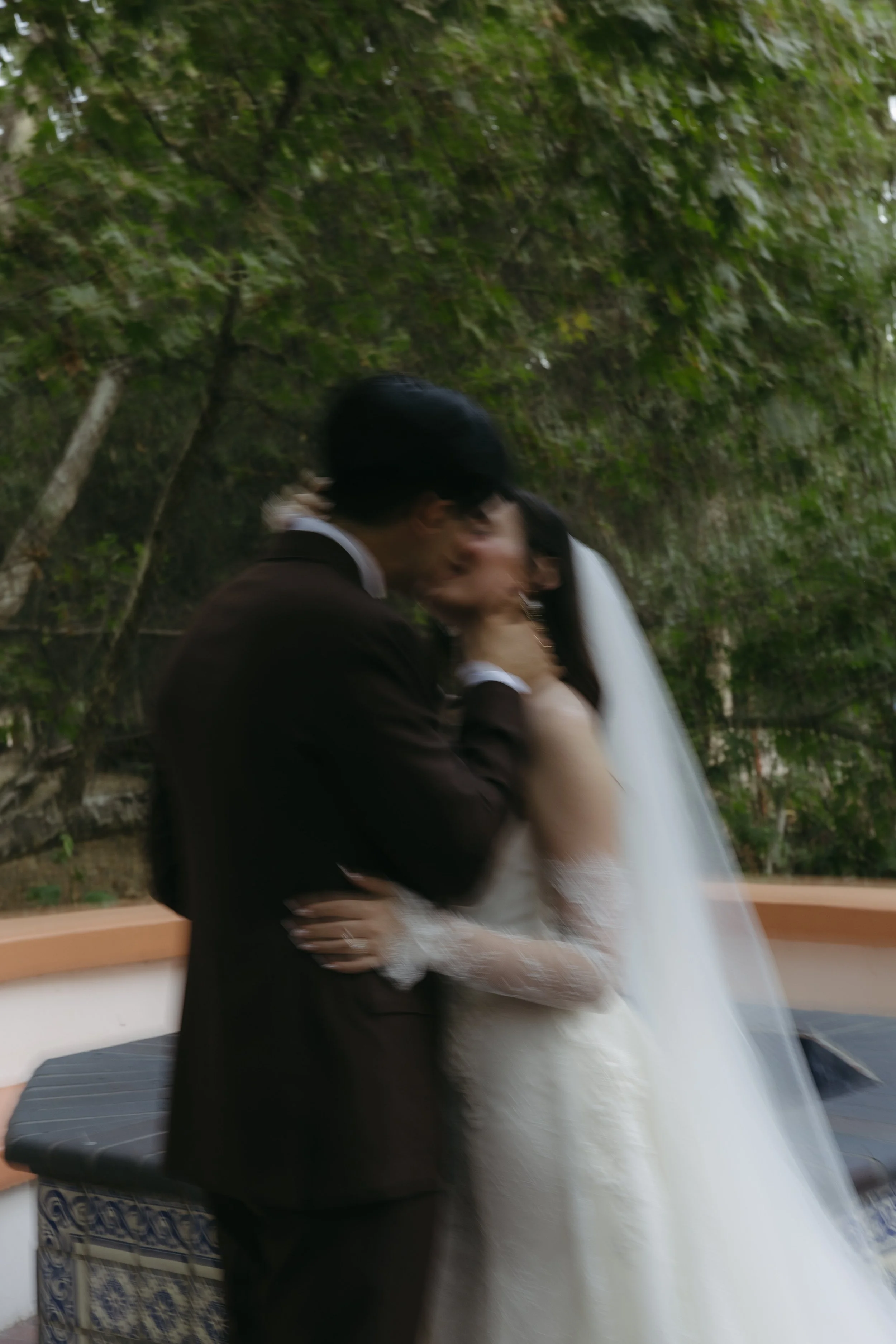 Blurred photo of a bride and groom sharing a kiss outdoors under trees.