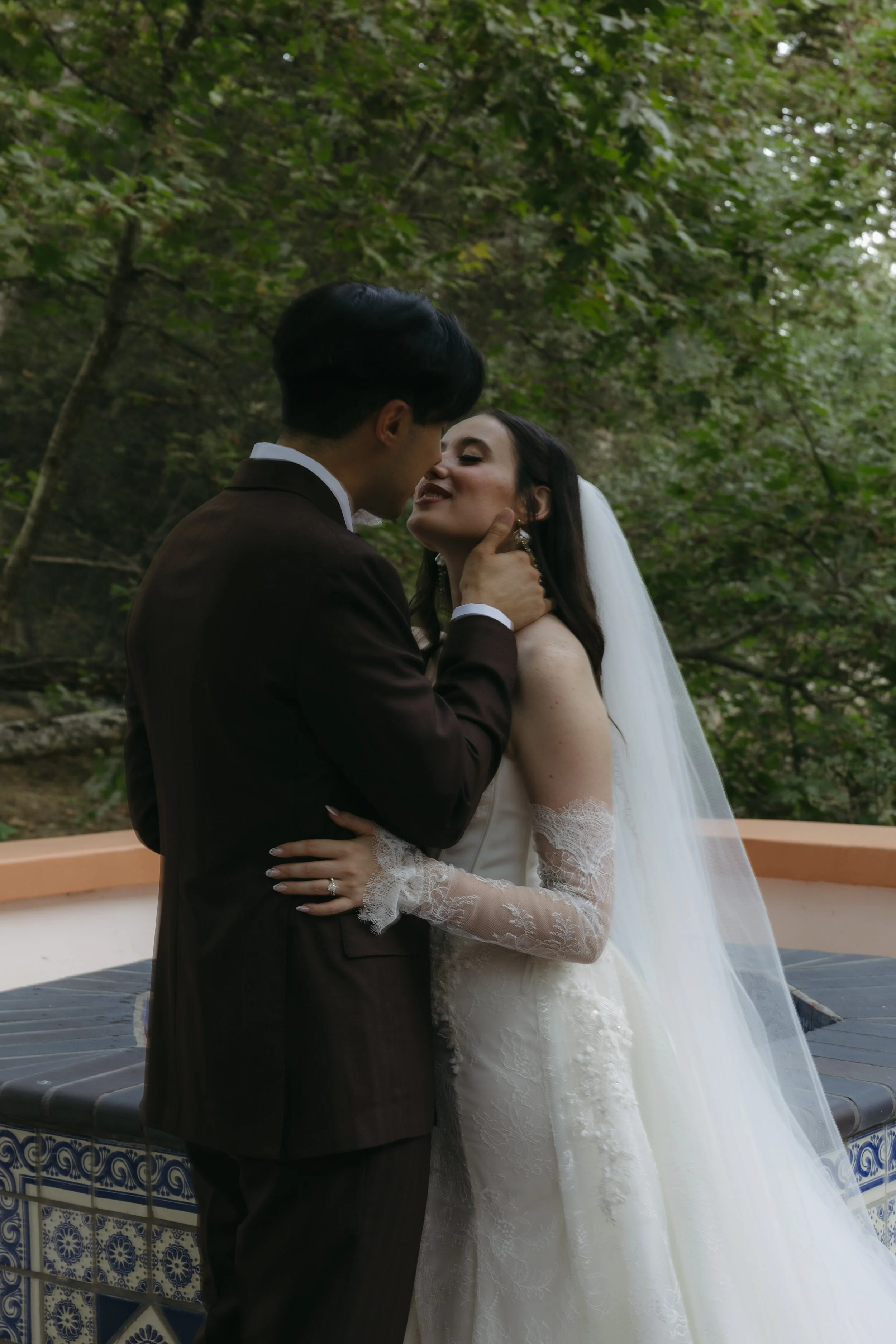 A newlywed couple sharing a romantic moment outdoors, surrounded by trees, with the bride wearing a wedding dress and veil, and the groom in a dark brown suit.