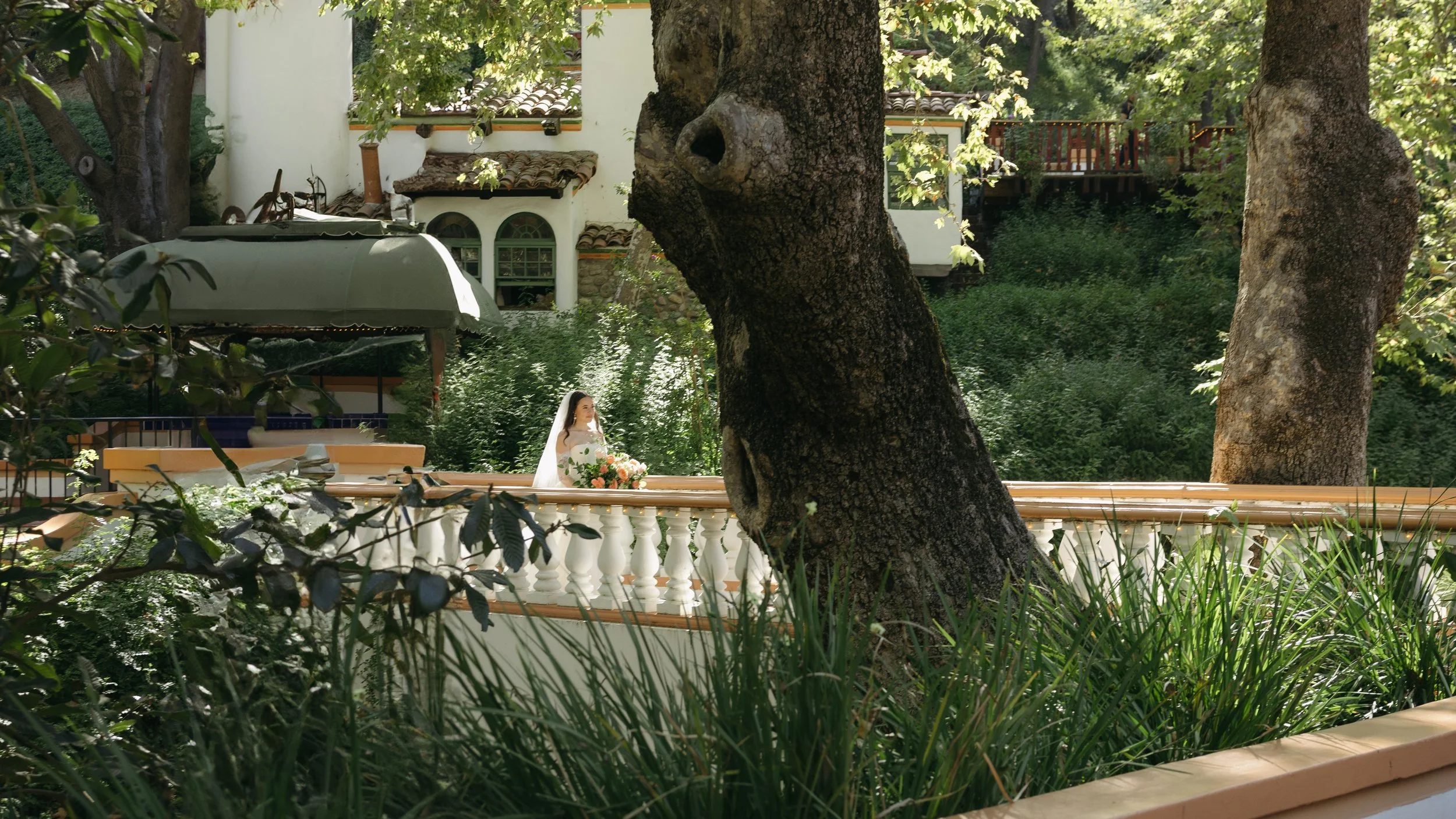 A bride holding a bouquet of flowers standing by a decorative white balcony in a lush green garden with large trees and a white house in the background at a Rancho Las Lomas Wedding in Los Angeles
